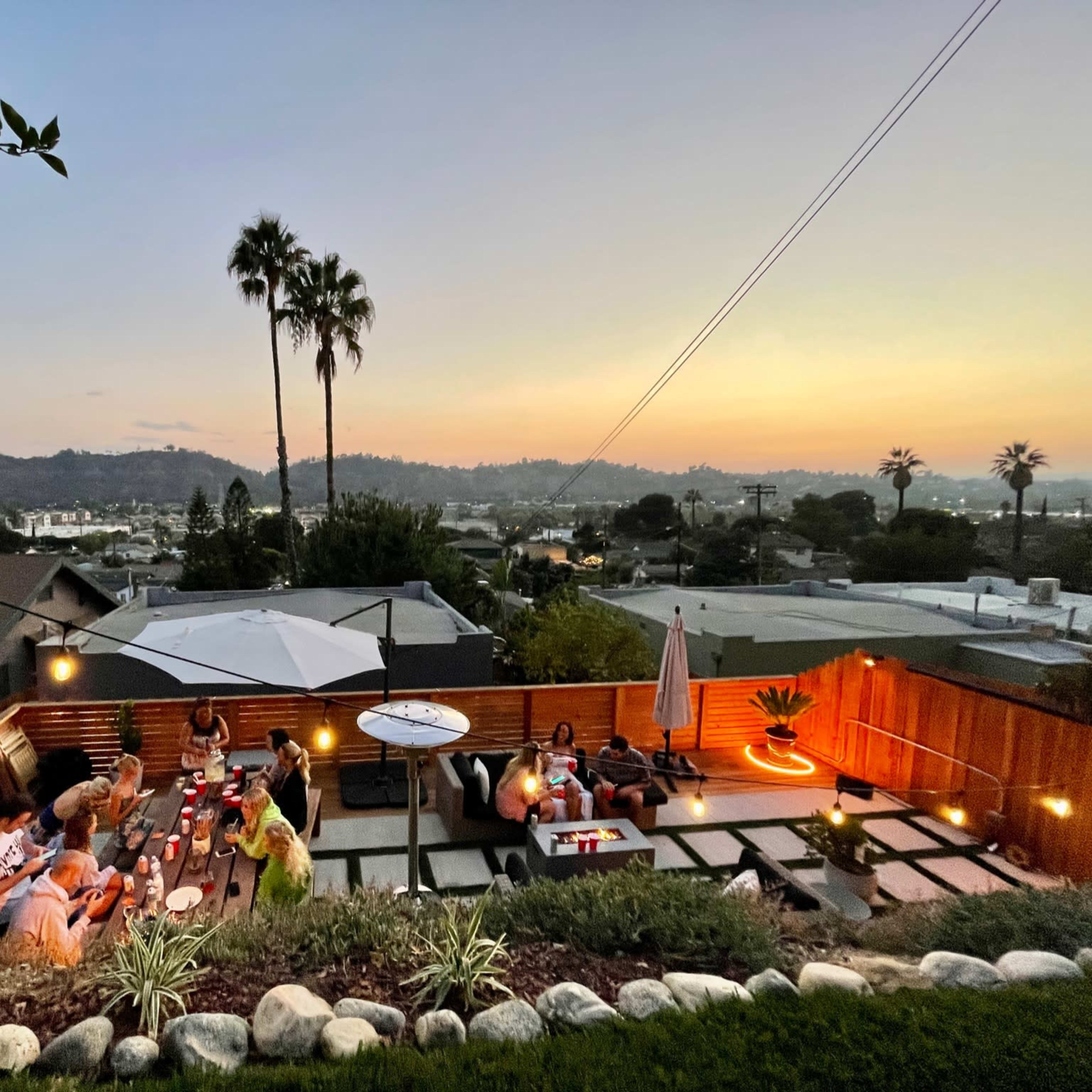 A large group of people gathers for an evening dinner on a hillside patio, with city lights and mountains visible in the background at sunset.