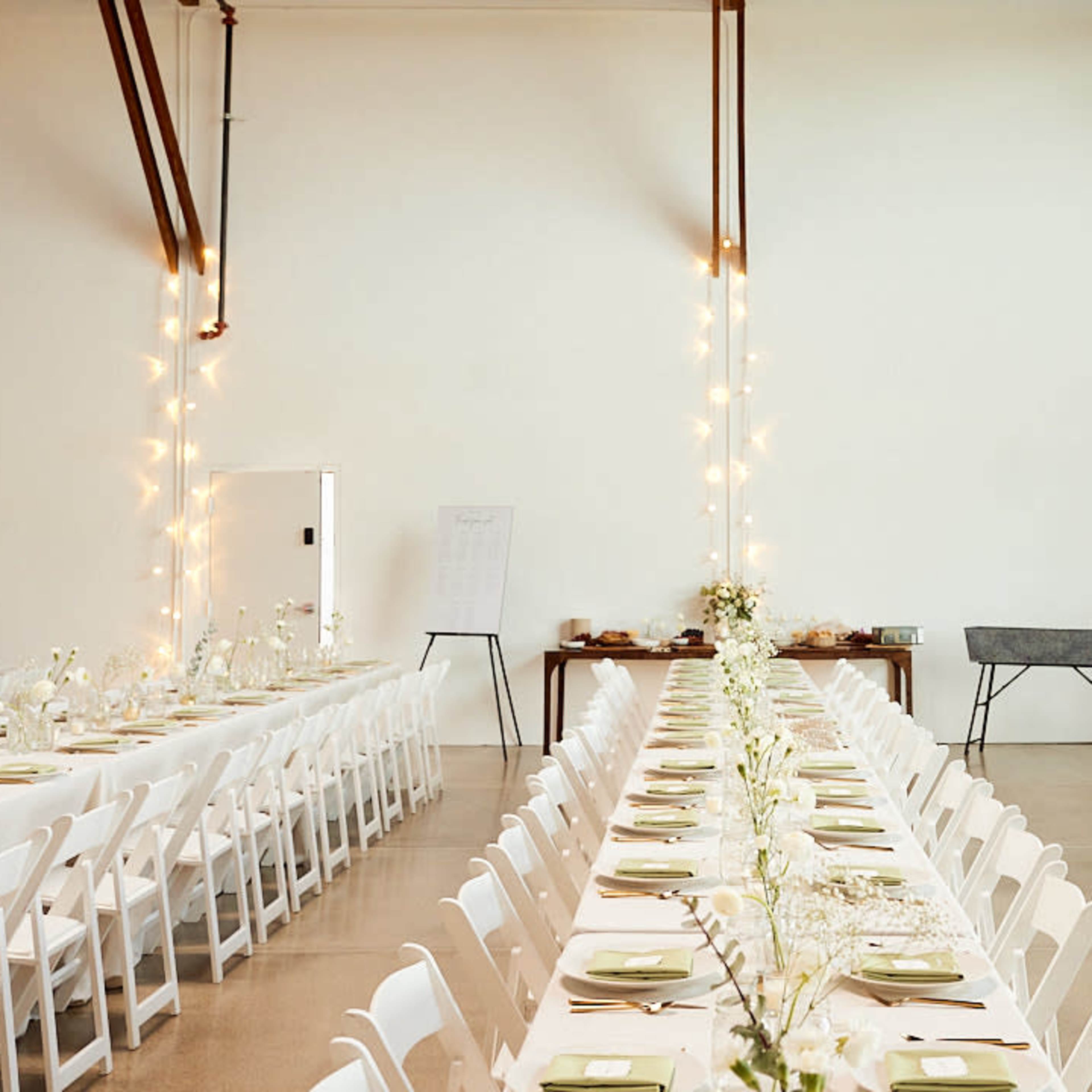 A spacious venue features a long table set with white chairs and floral centerpieces, illuminated by soft string lights along the walls.