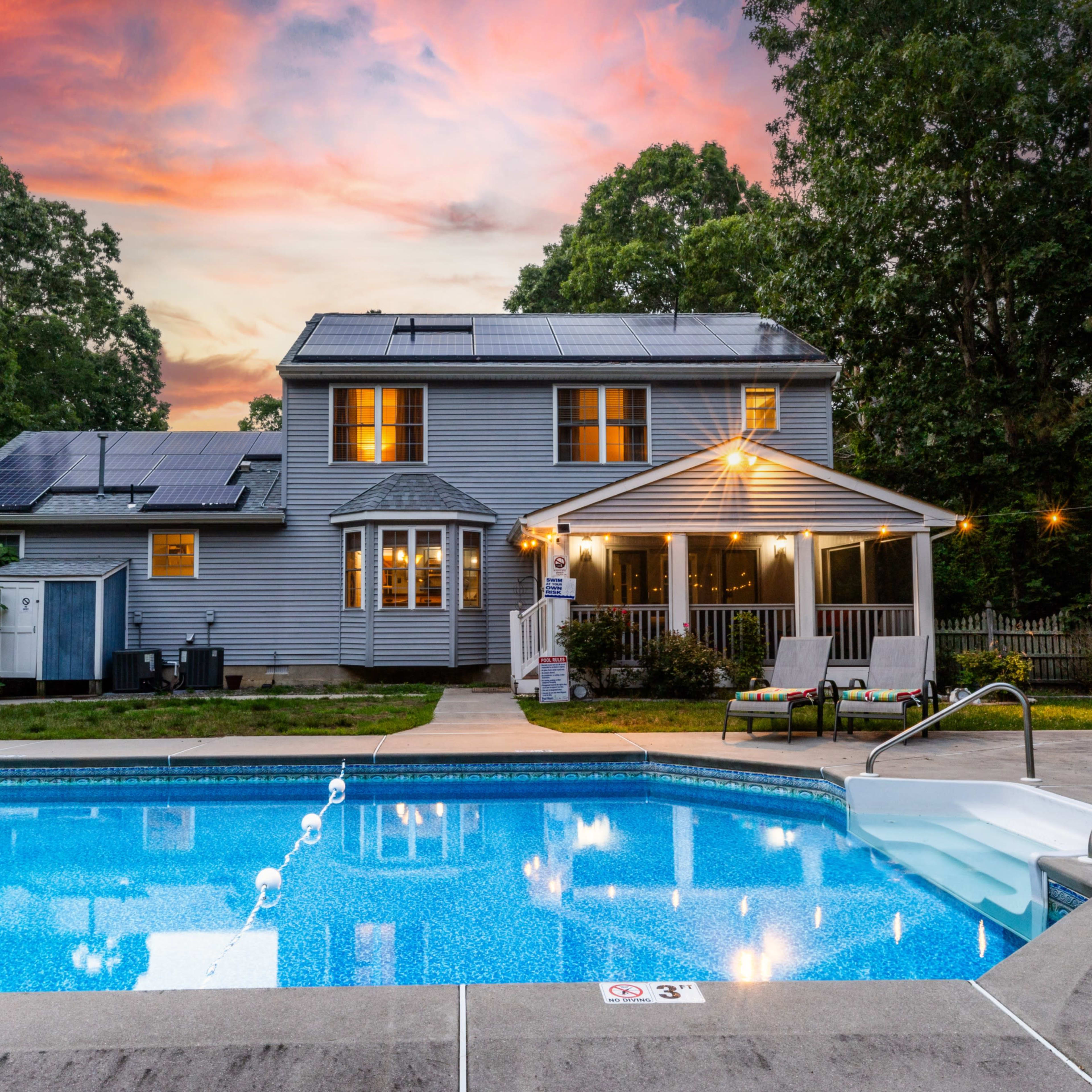 A two-story house with solar panels and a pool in the foreground, set against a colorful sunset sky.