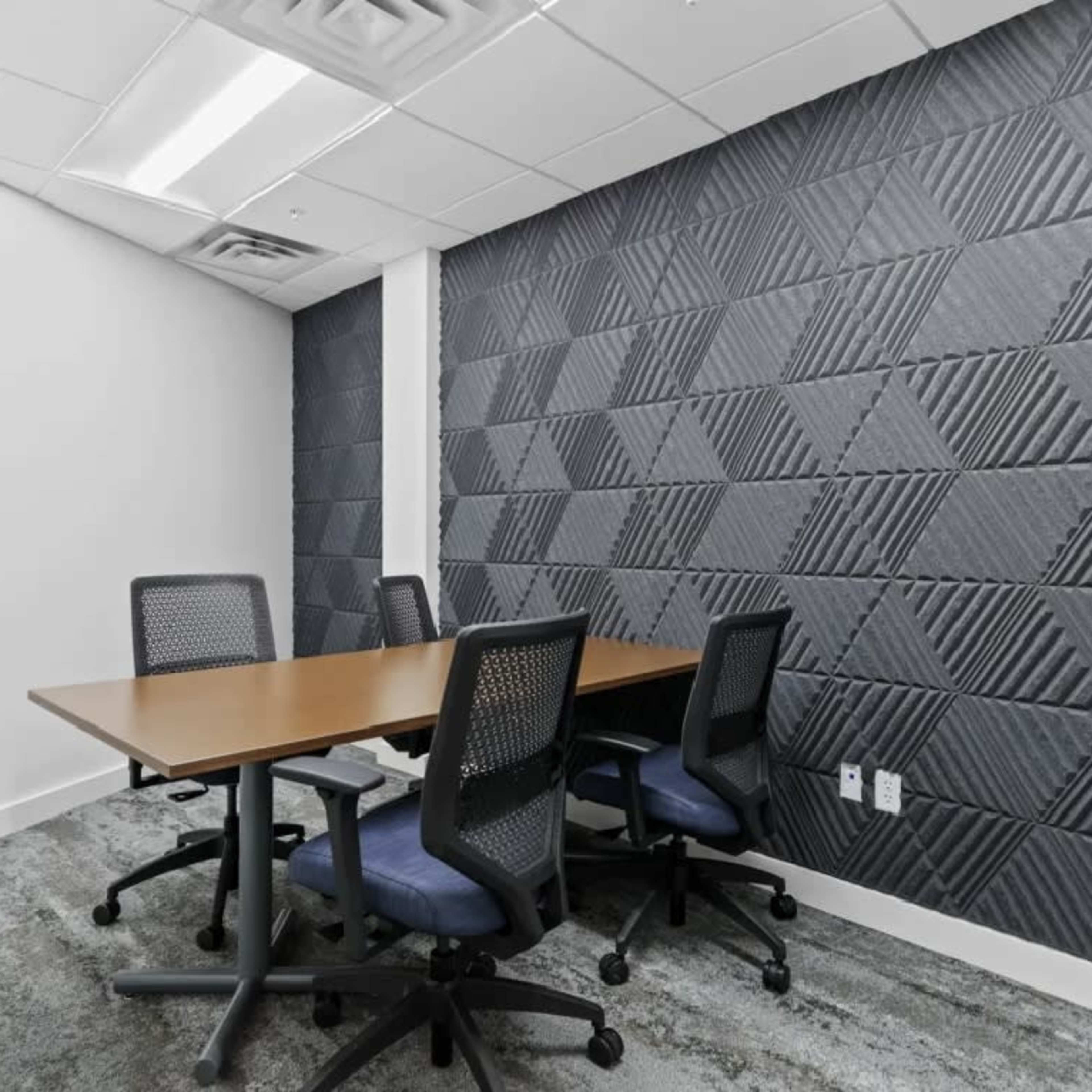 A small conference room features a wooden table surrounded by four office chairs, with textured gray walls and carpeted flooring.