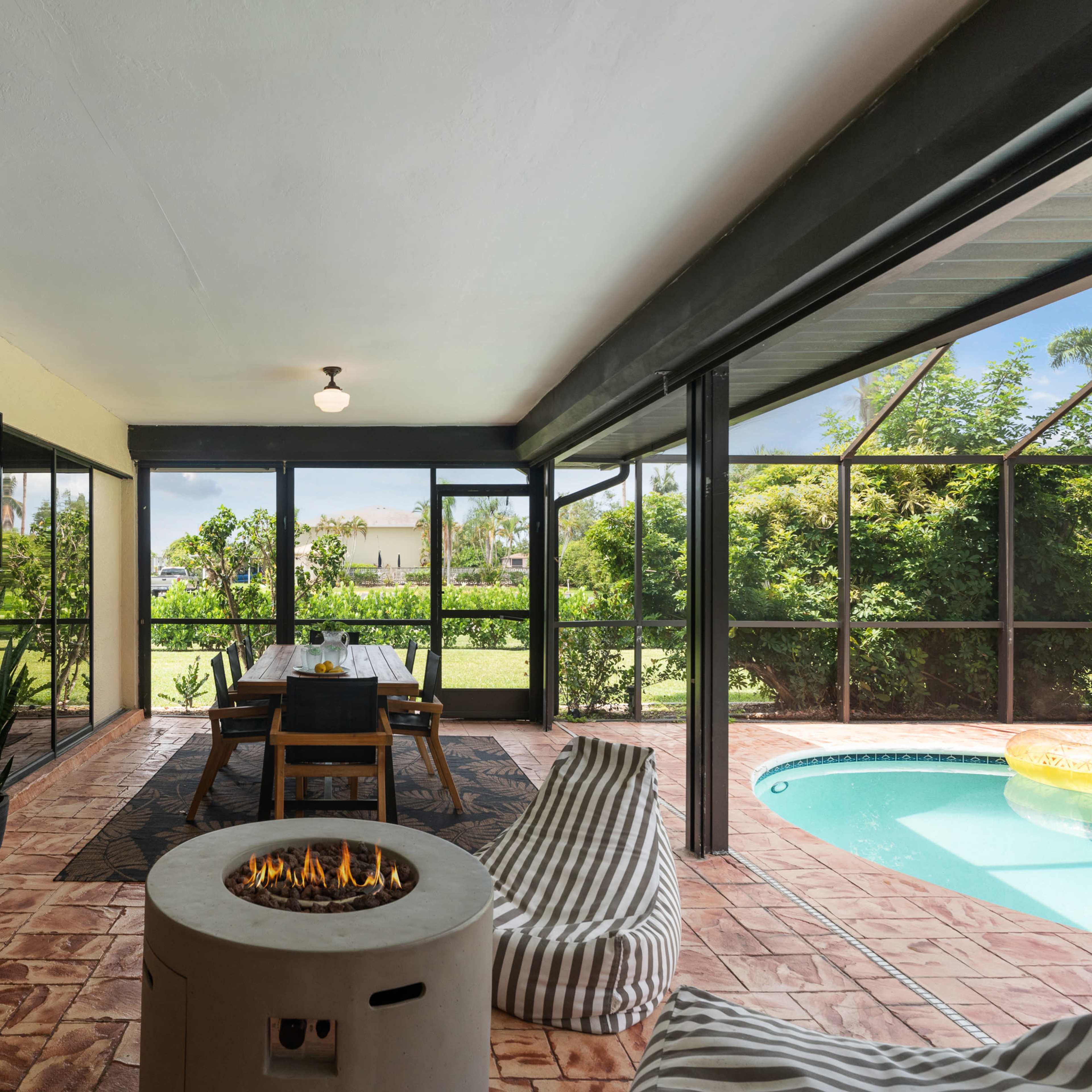 A screened patio with a dining table, a circular fire pit, a swimming pool, and lush greenery in the background.