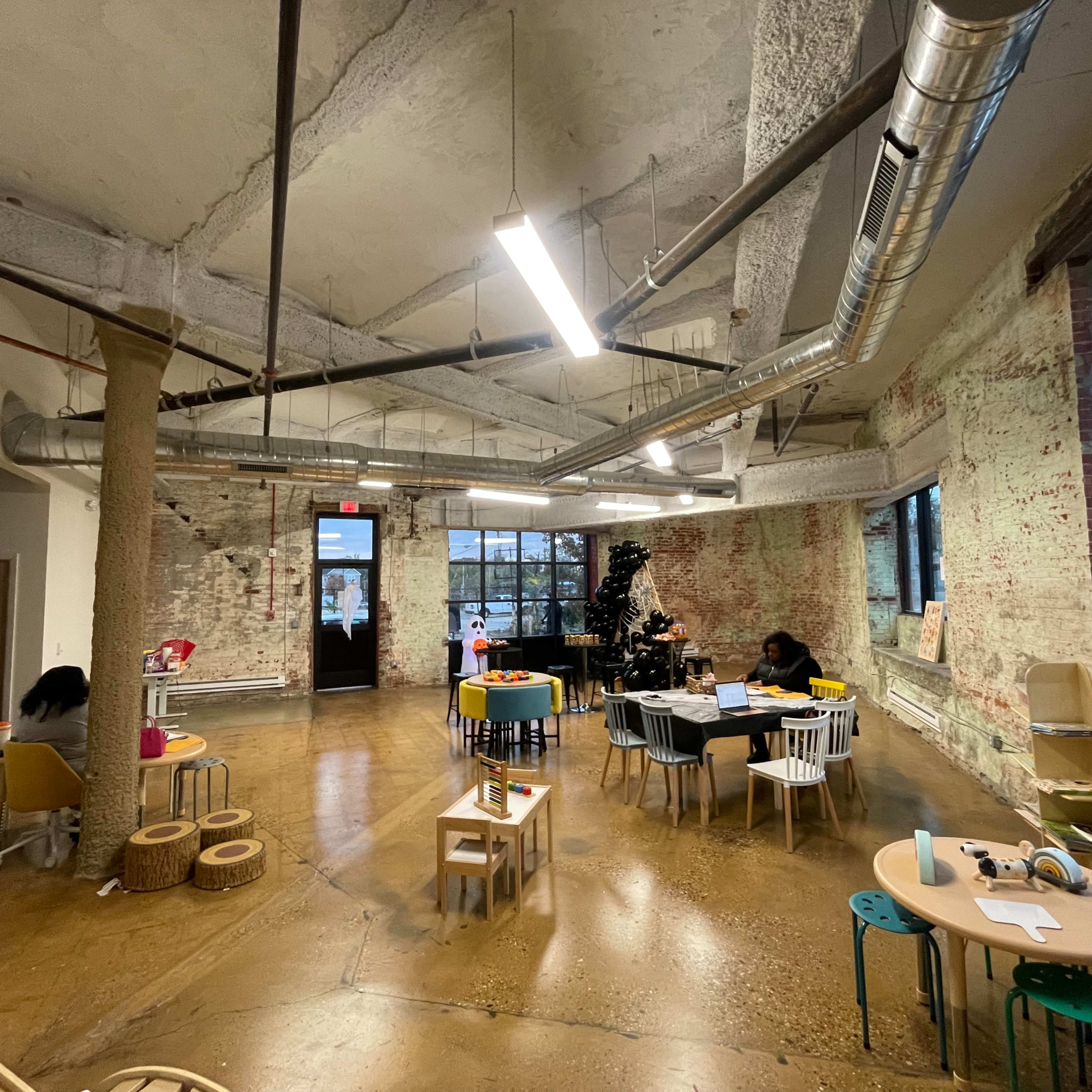 An open play area with various child-sized tables and chairs, exposed brick walls, and large windows providing natural light.