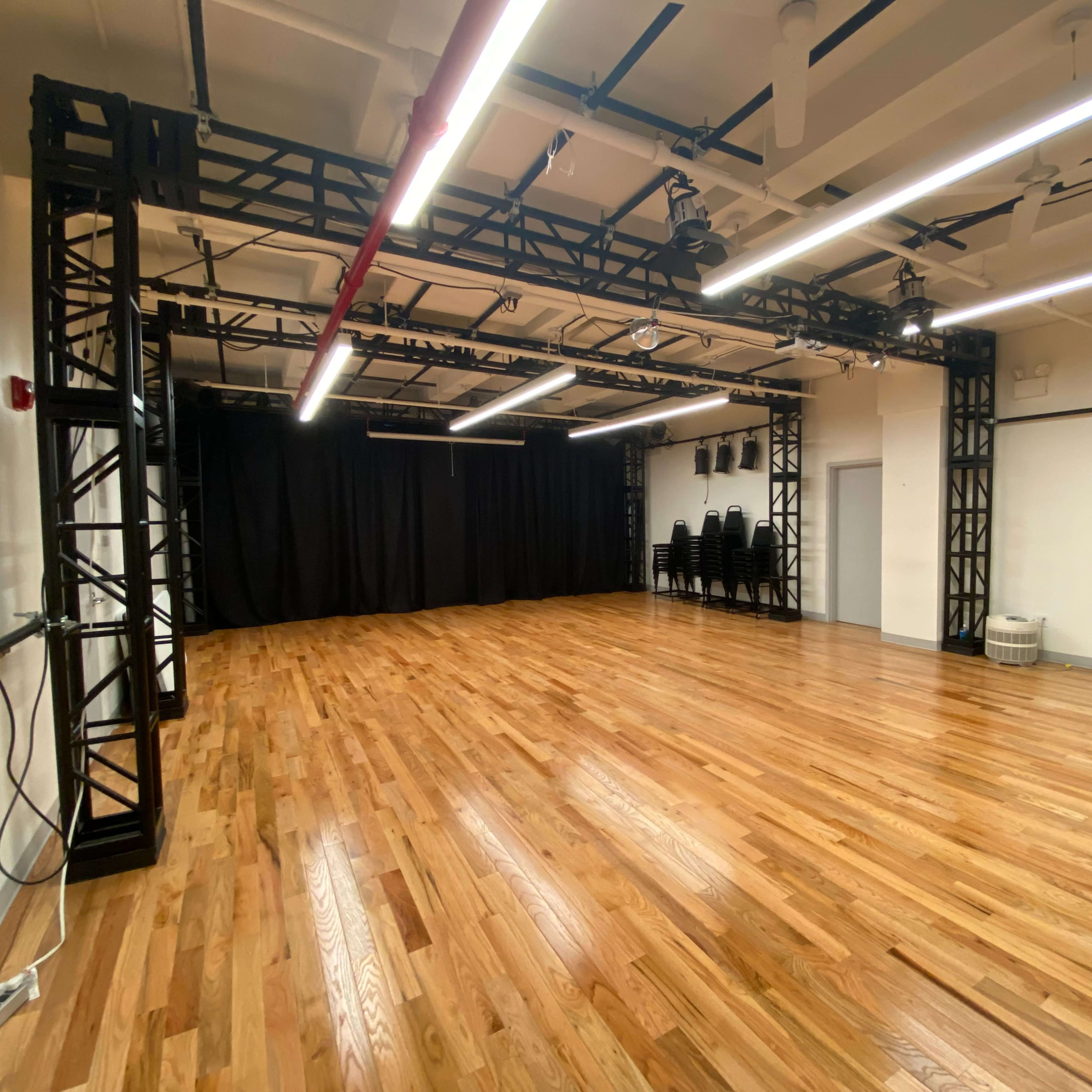 A spacious rehearsal studio with polished wooden flooring, black fabric backdrop, and a grid of overhead lights.
