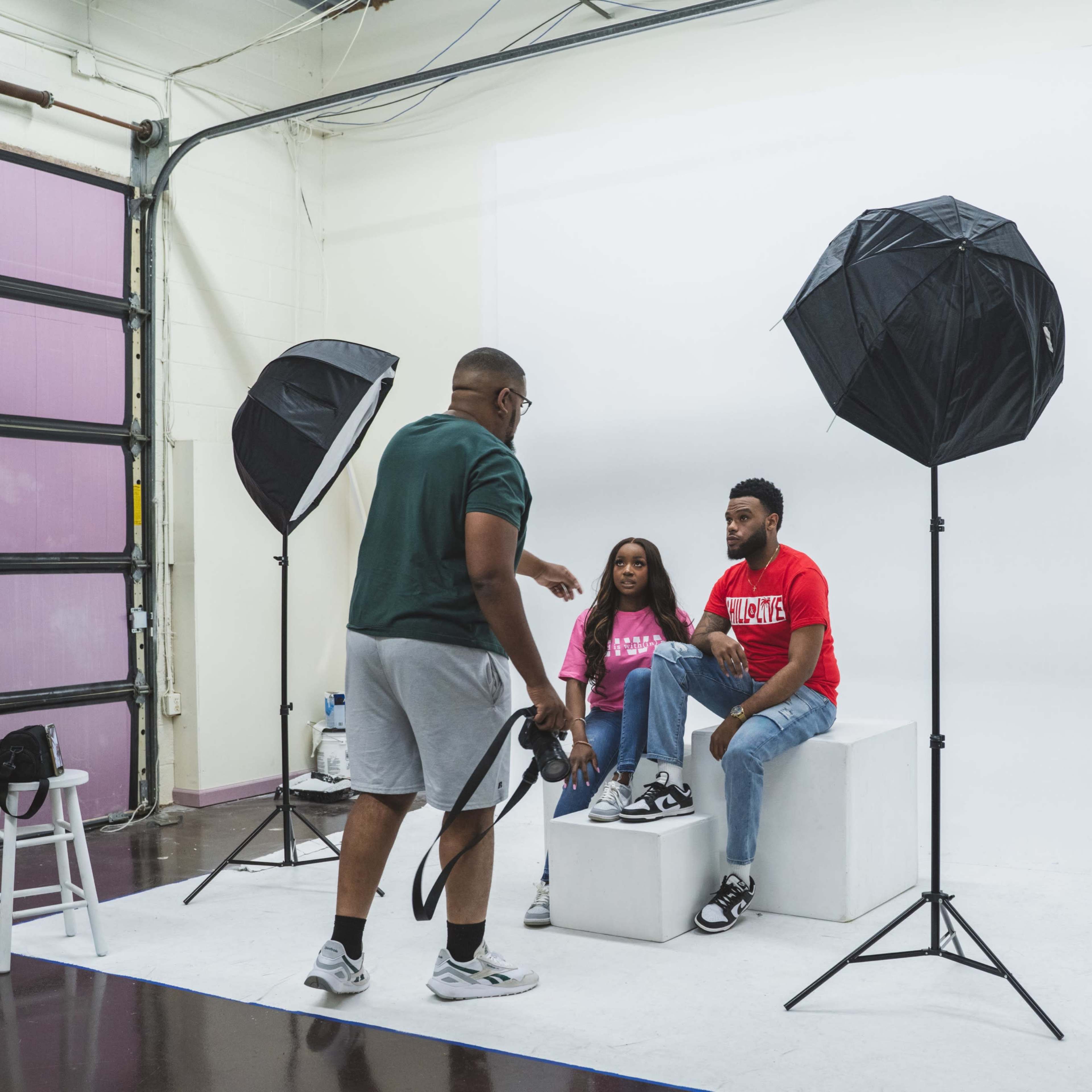 A photographer directs a couple posing on a pedestal in a studio with softbox lights.