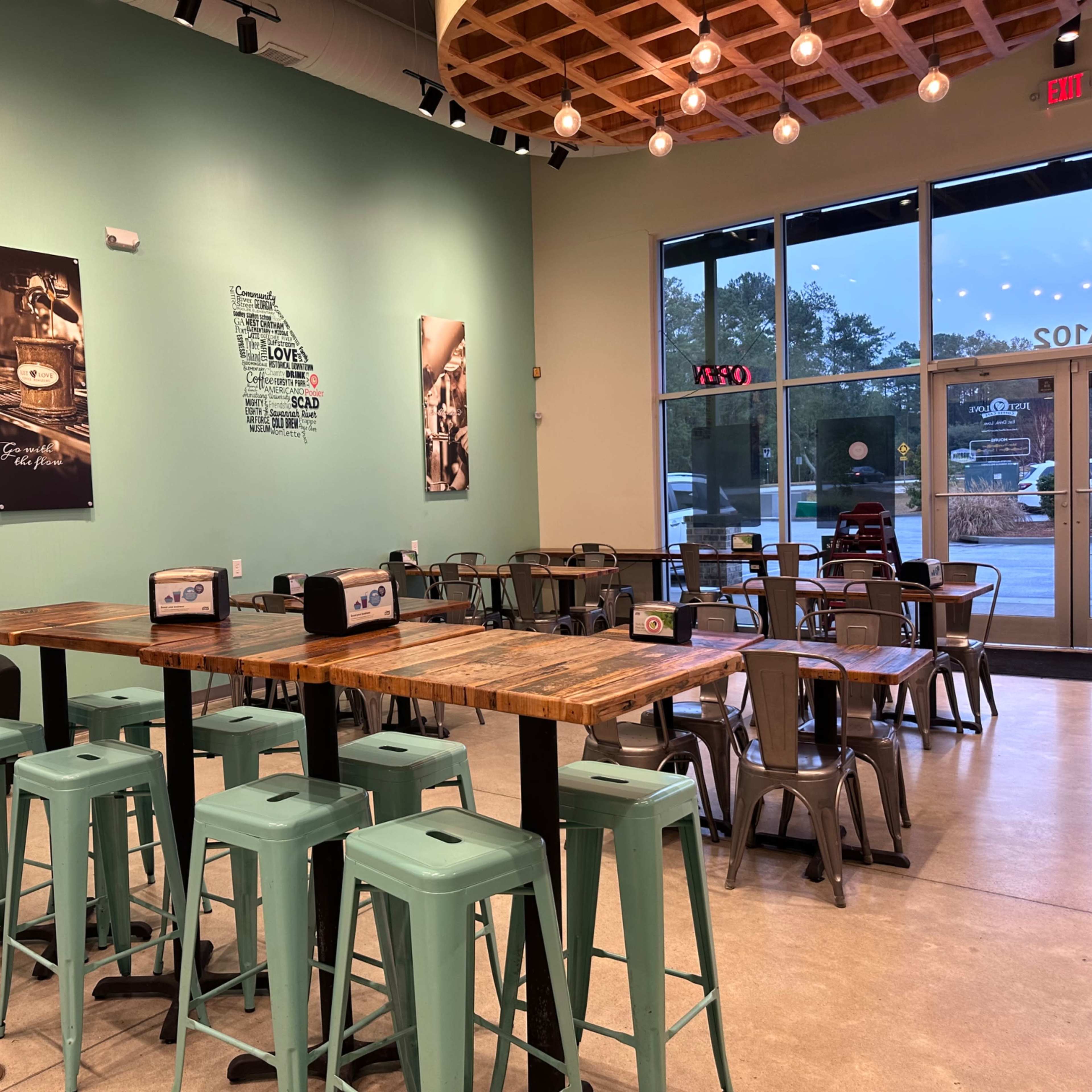 A cozy café interior with metal stools and wooden tables, featuring large windows and wall art.