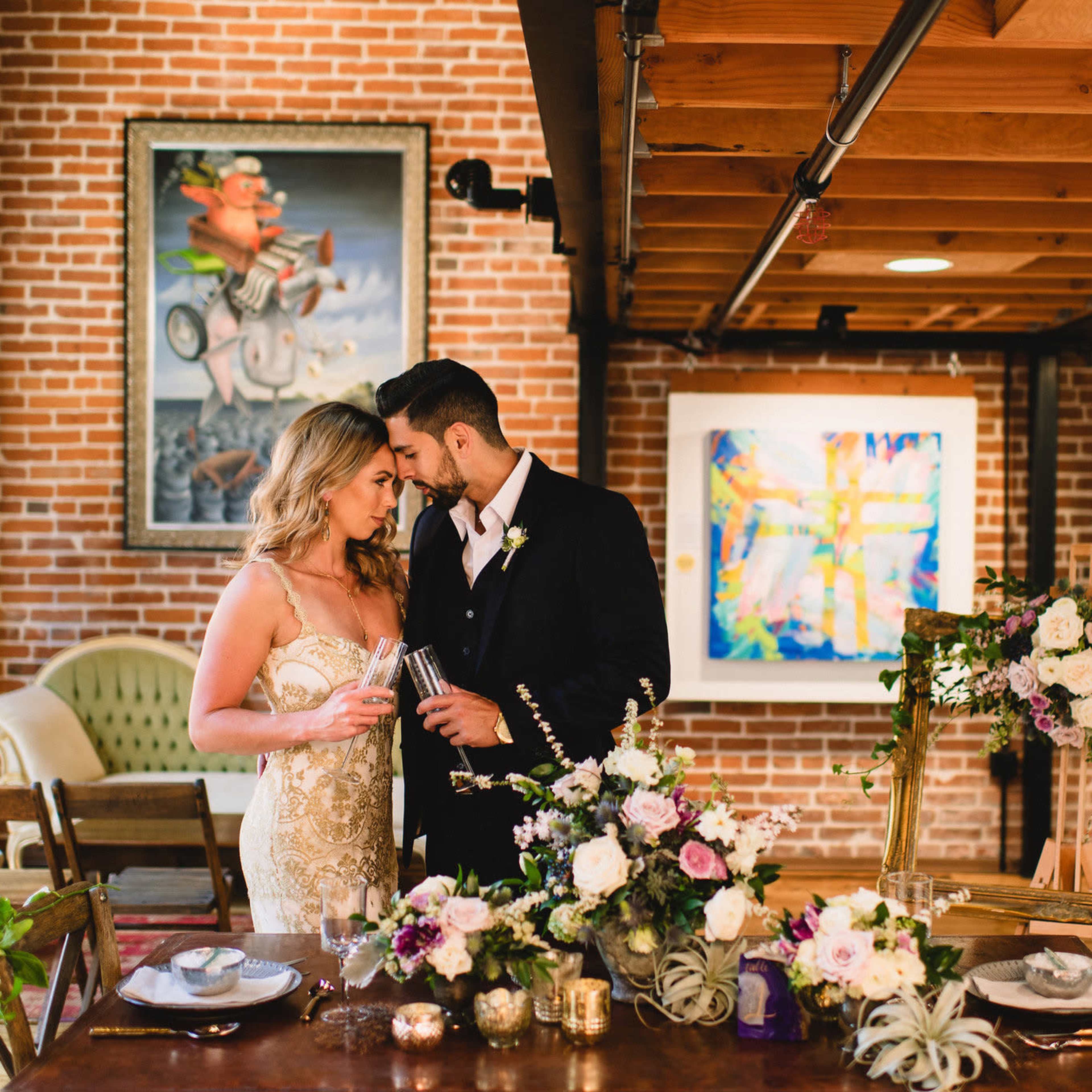 A couple stands together at a beautifully arranged table covered with flowers and dinnerware, set against a backdrop of exposed brick walls and artwork.