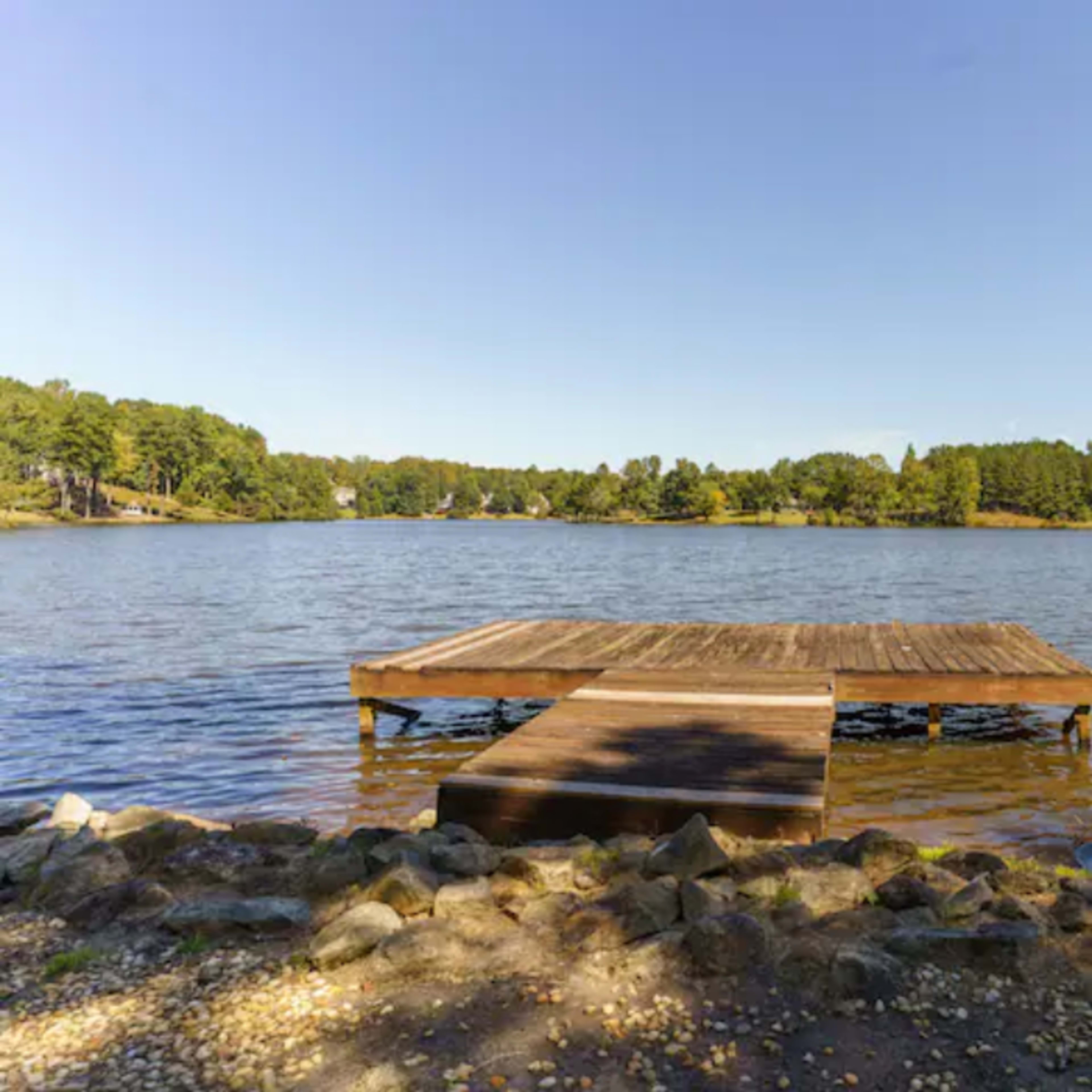 A wooden dock extends into a calm lake surrounded by trees under a clear blue sky.