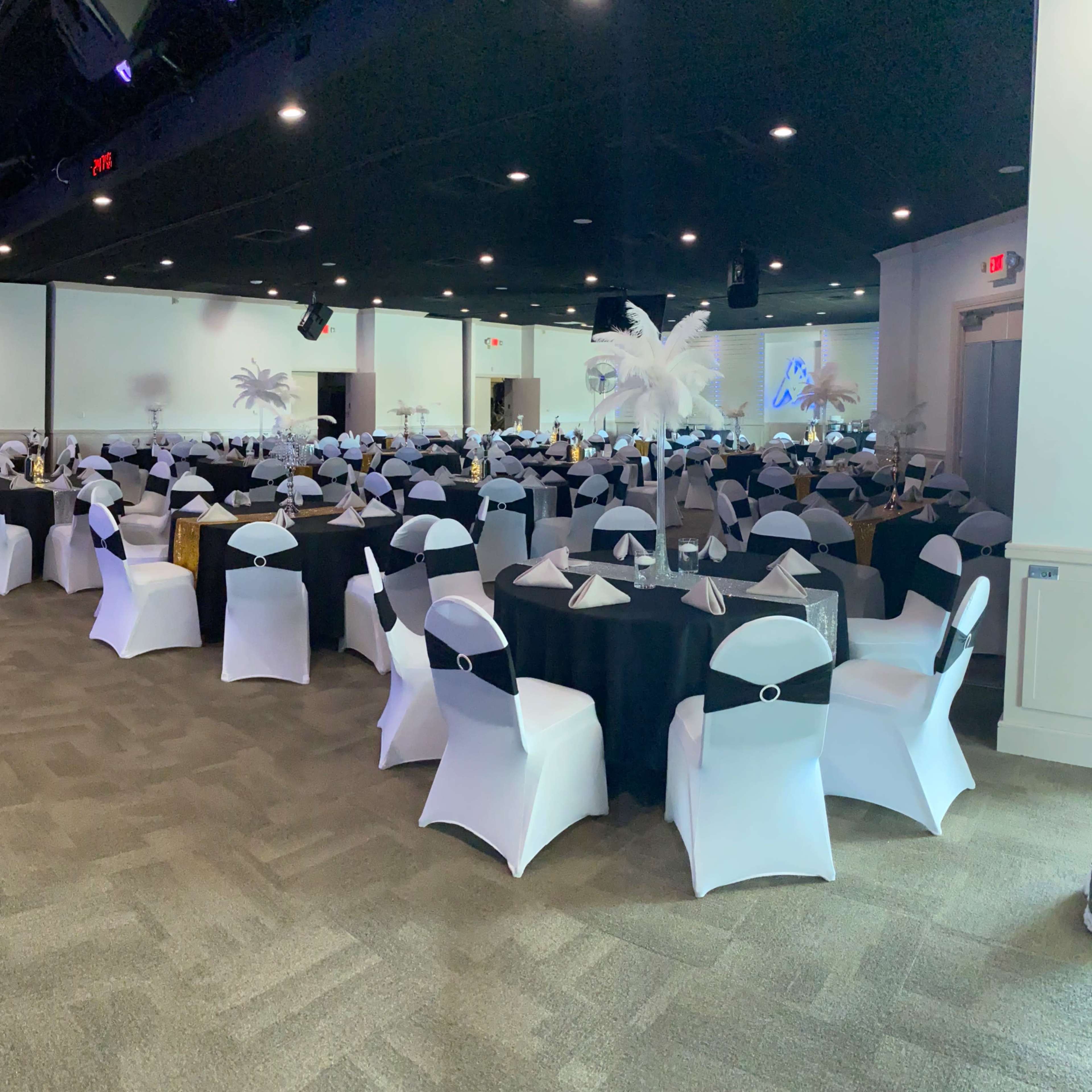 A spacious banquet hall is set up with round tables covered in black and white tablecloths, featuring neatly arranged place settings and decorative centerpieces.