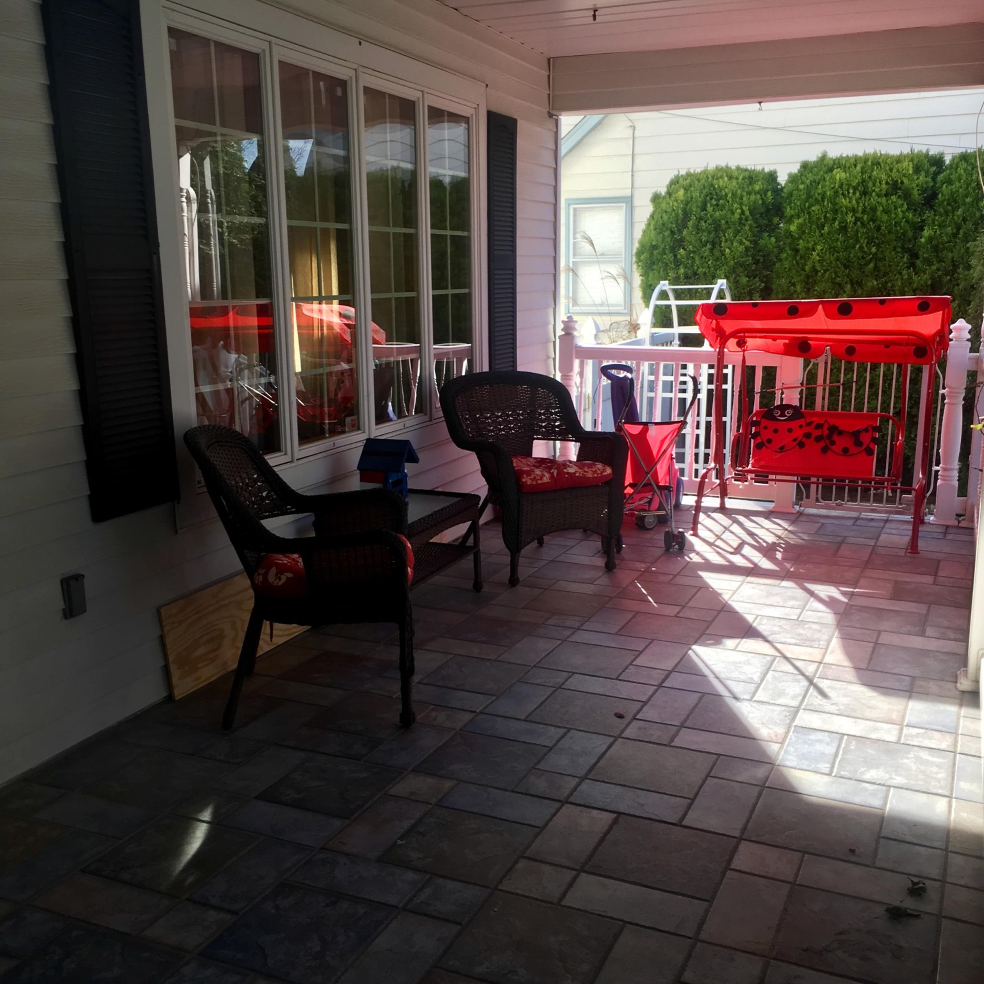 A covered porch with two wicker chairs, a small table, and a red cart, all set on a stone-tiled floor.