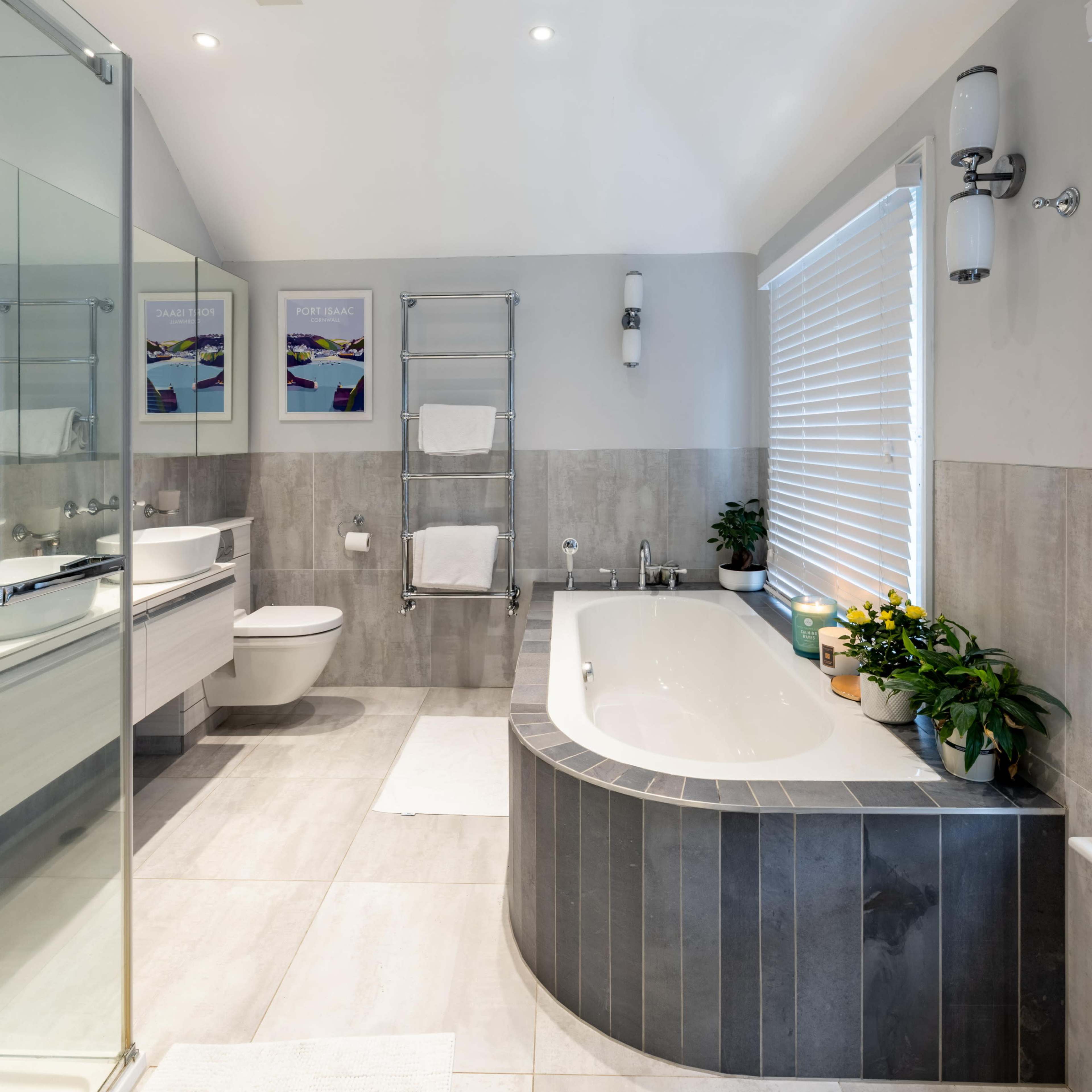 A modern bathroom featuring a curved bathtub, a glass shower enclosure, a toilet, and a sink area with plants and towels.