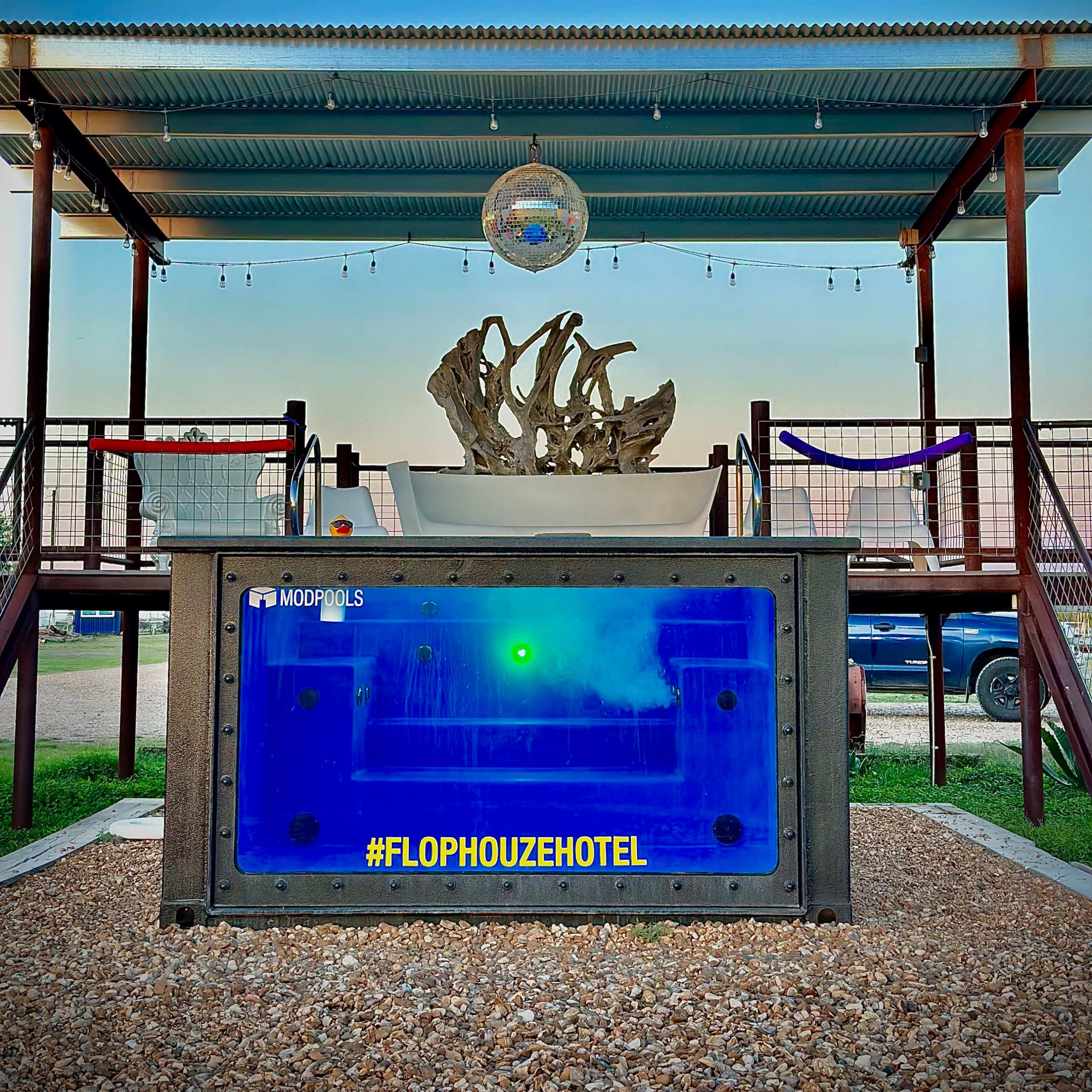 A unique hotel setup featuring a large, illuminated blue structure under a metal roof, with a decorative piece above it and a hashtag, "#FLOPHOUZEHOTEL," prominently displayed.