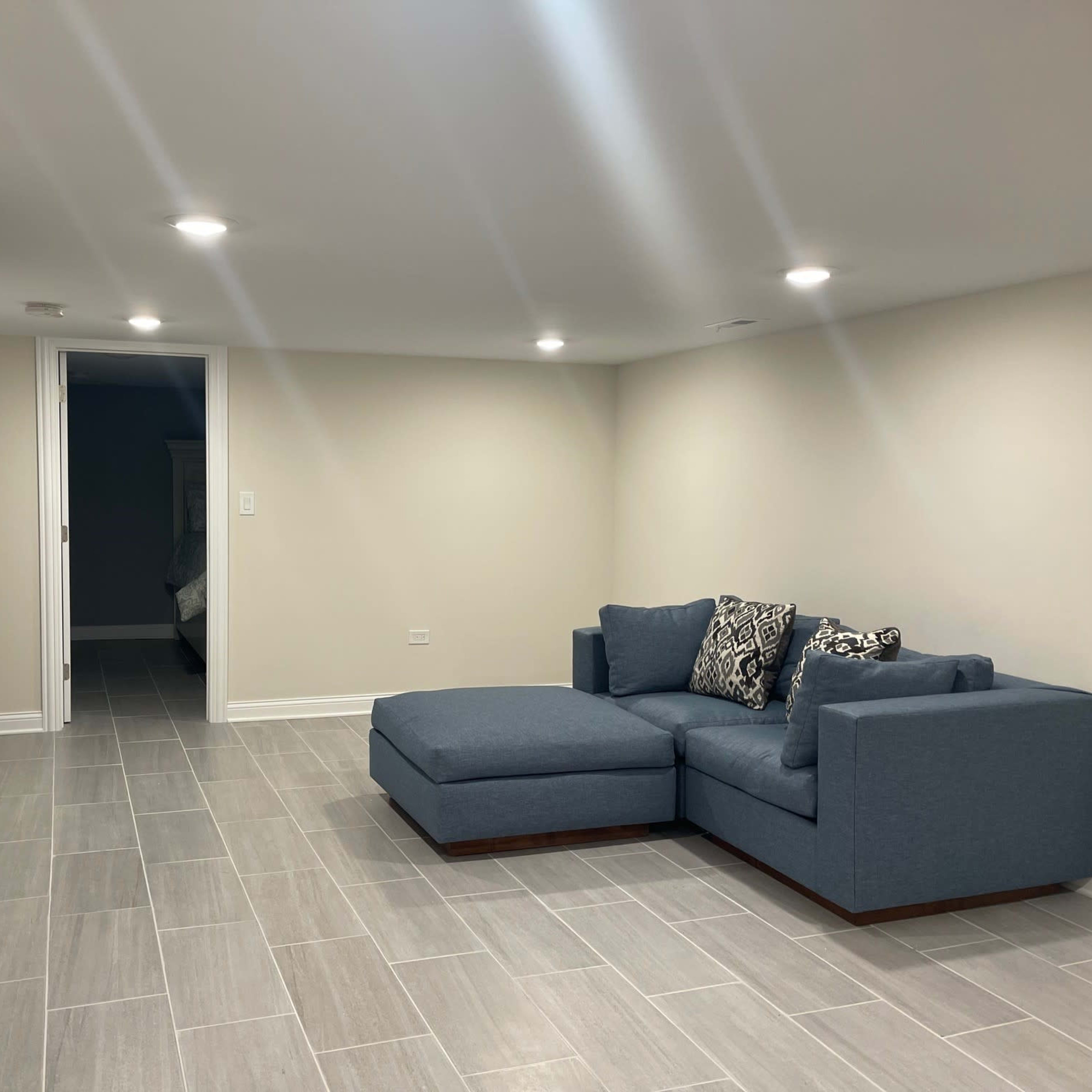 A finished basement with gray tile flooring, a blue sectional couch, and a wall-mounted television.
