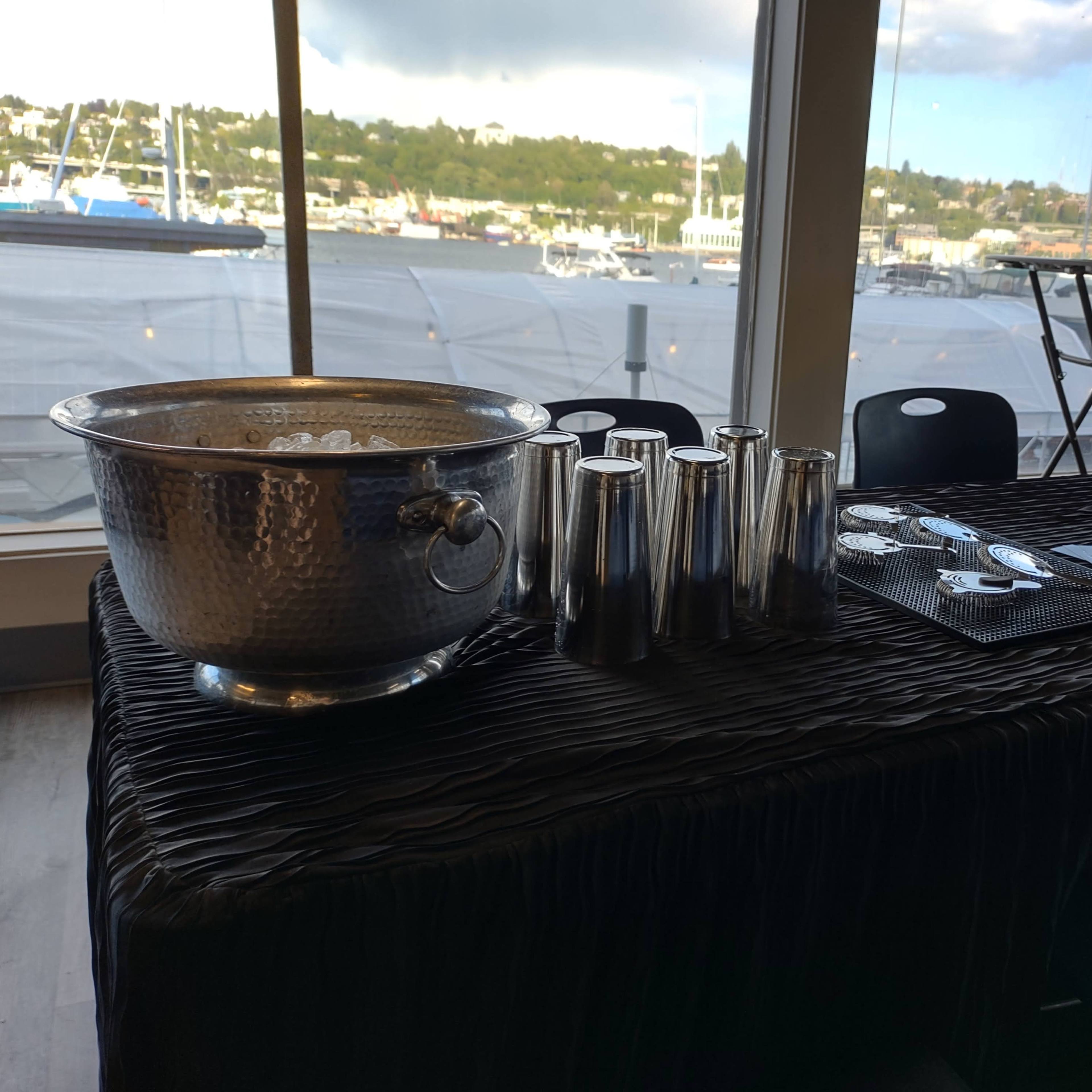 A table is set up near a large window, displaying a silver ice bucket, cocktail shakers, and a bowl of fruit, with a view of boats docked outside.