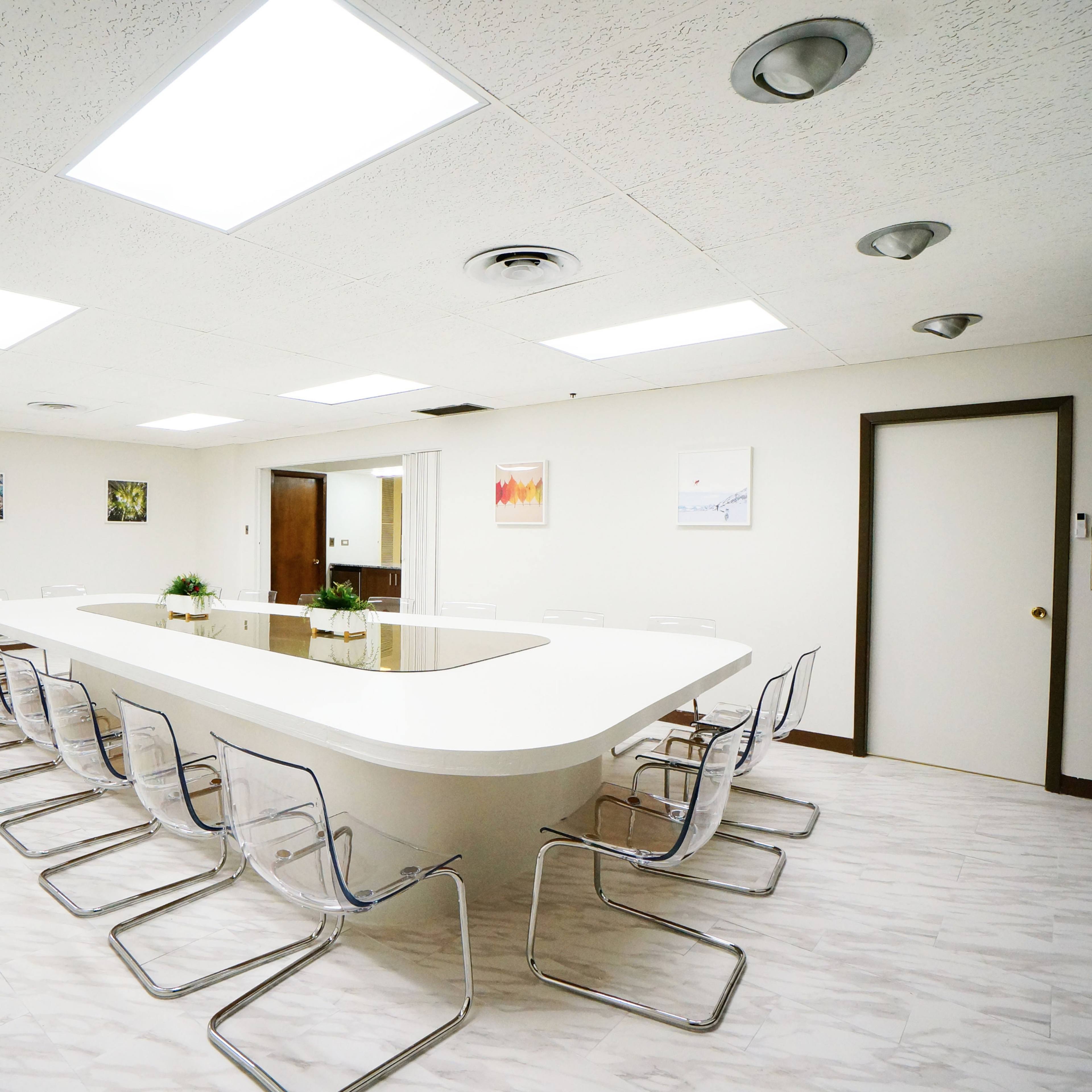 A modern conference room features a long white table surrounded by transparent chairs, with light fixtures illuminating the space.