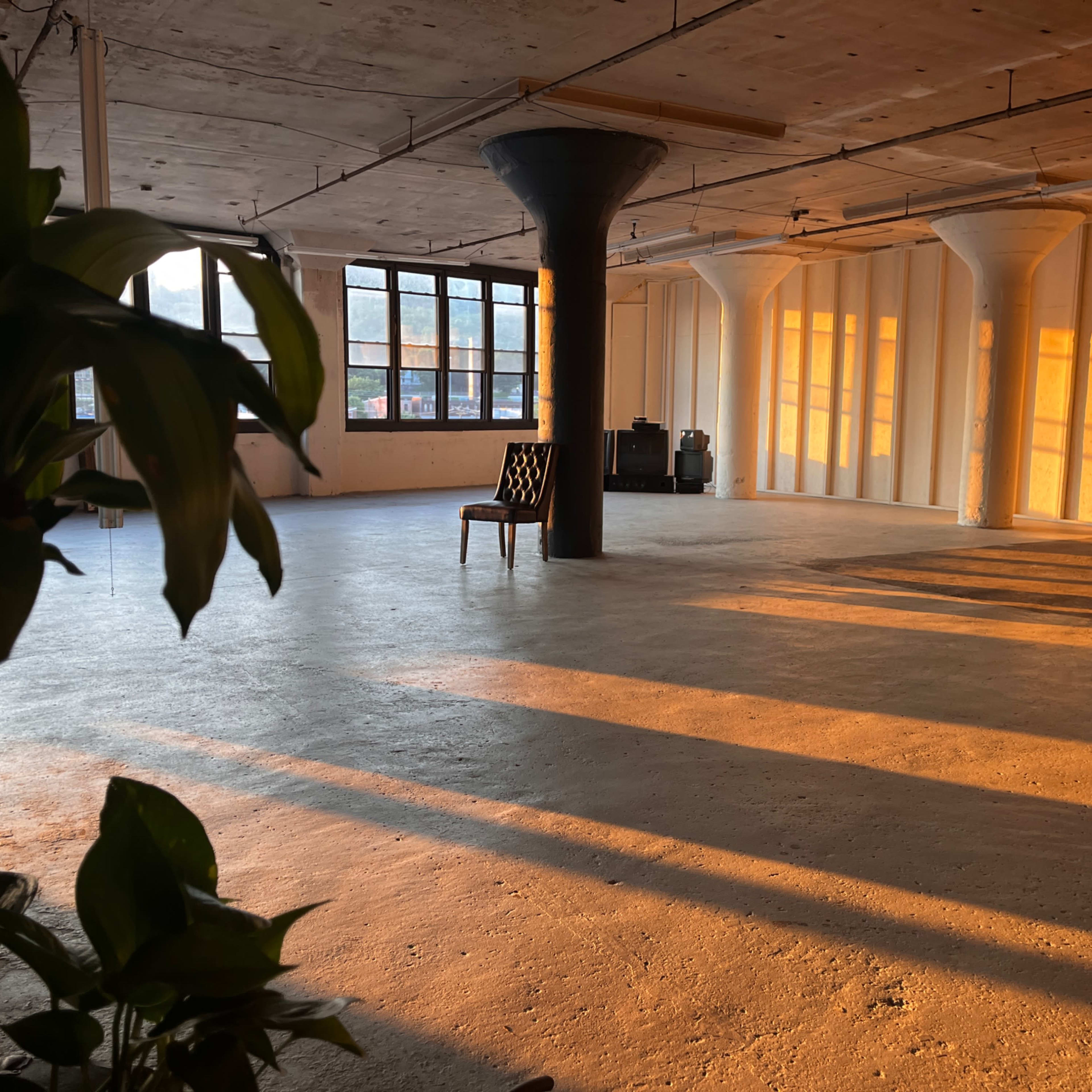 An empty industrial space with a single chair and long shadows cast by the setting sun through large windows.