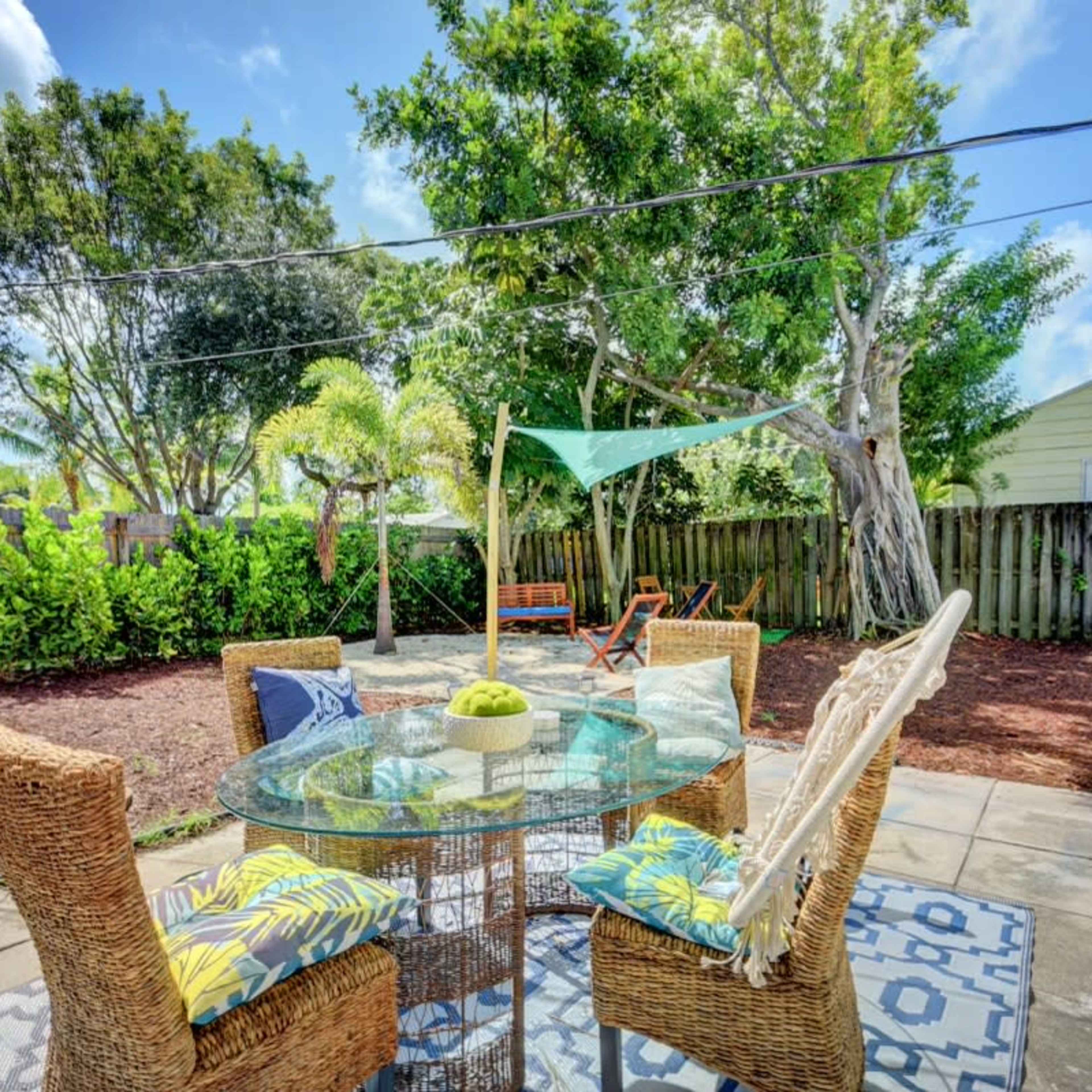 A patio area features a glass table surrounded by wicker chairs and decorative cushions, with greenery and a swing set in the background.