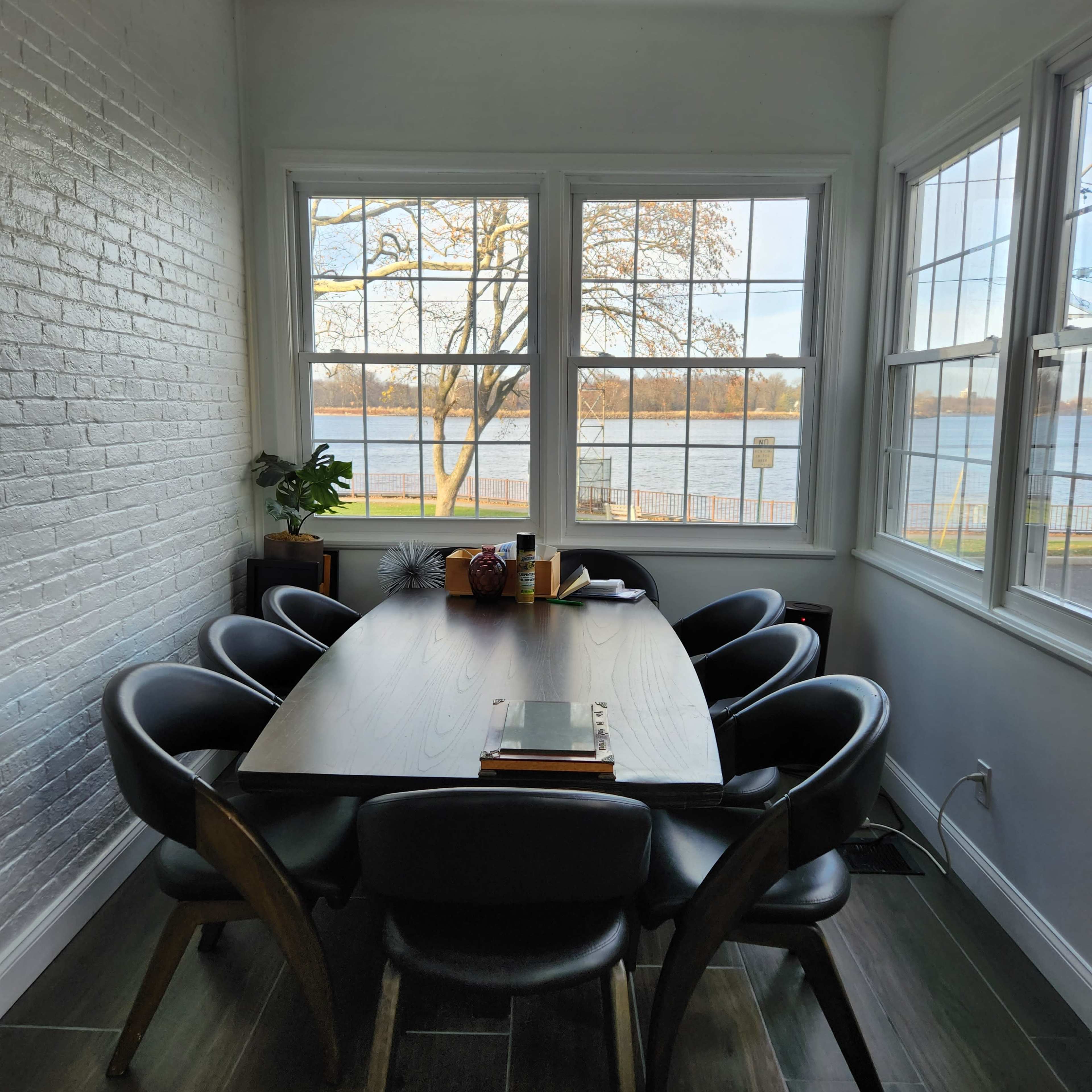 A long wooden dining table with six chairs is set against a large window overlooking a lake and trees.
