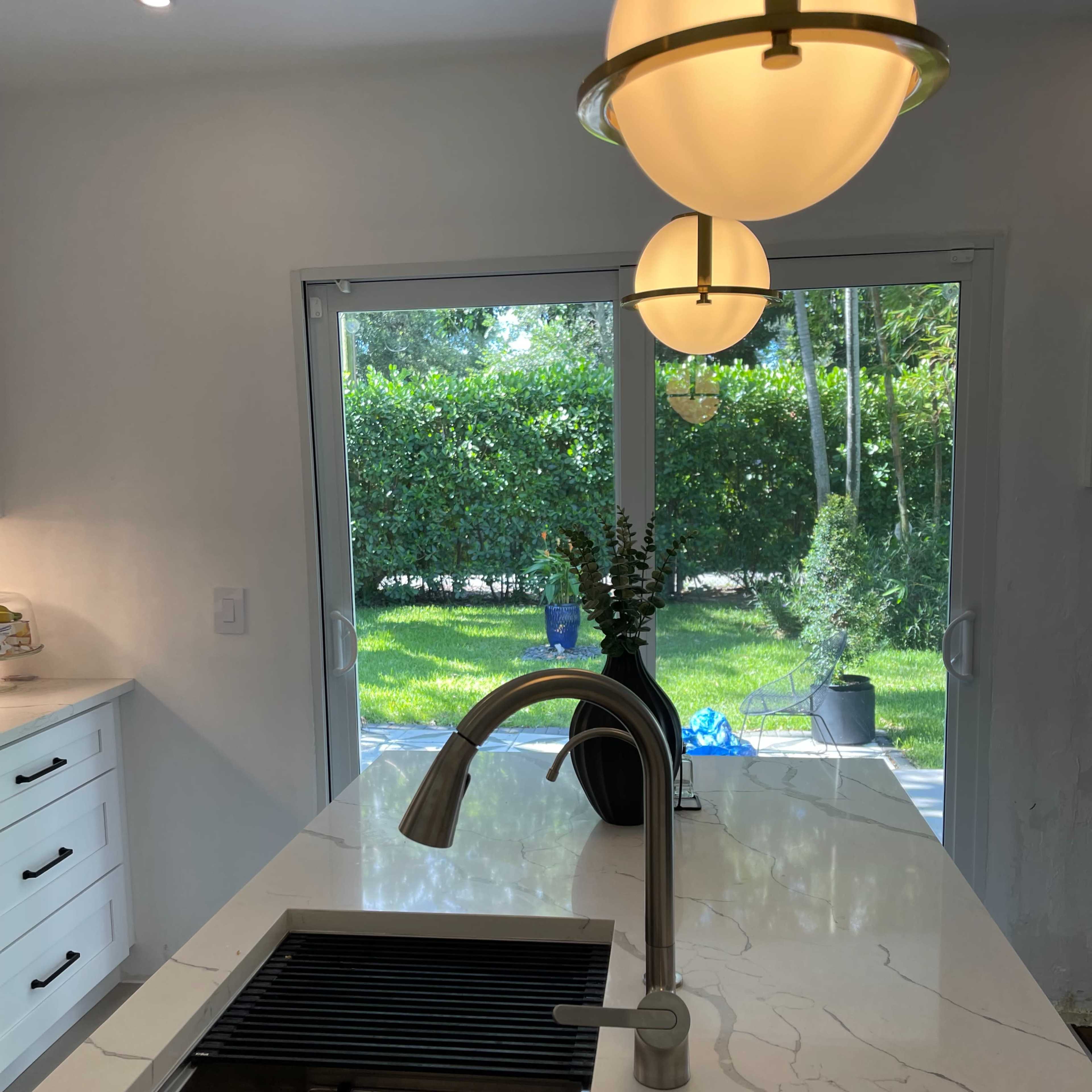 A modern kitchen features a sleek sink with a faucet, a marble countertop, and pendant lights, overlooking a garden through sliding glass doors.