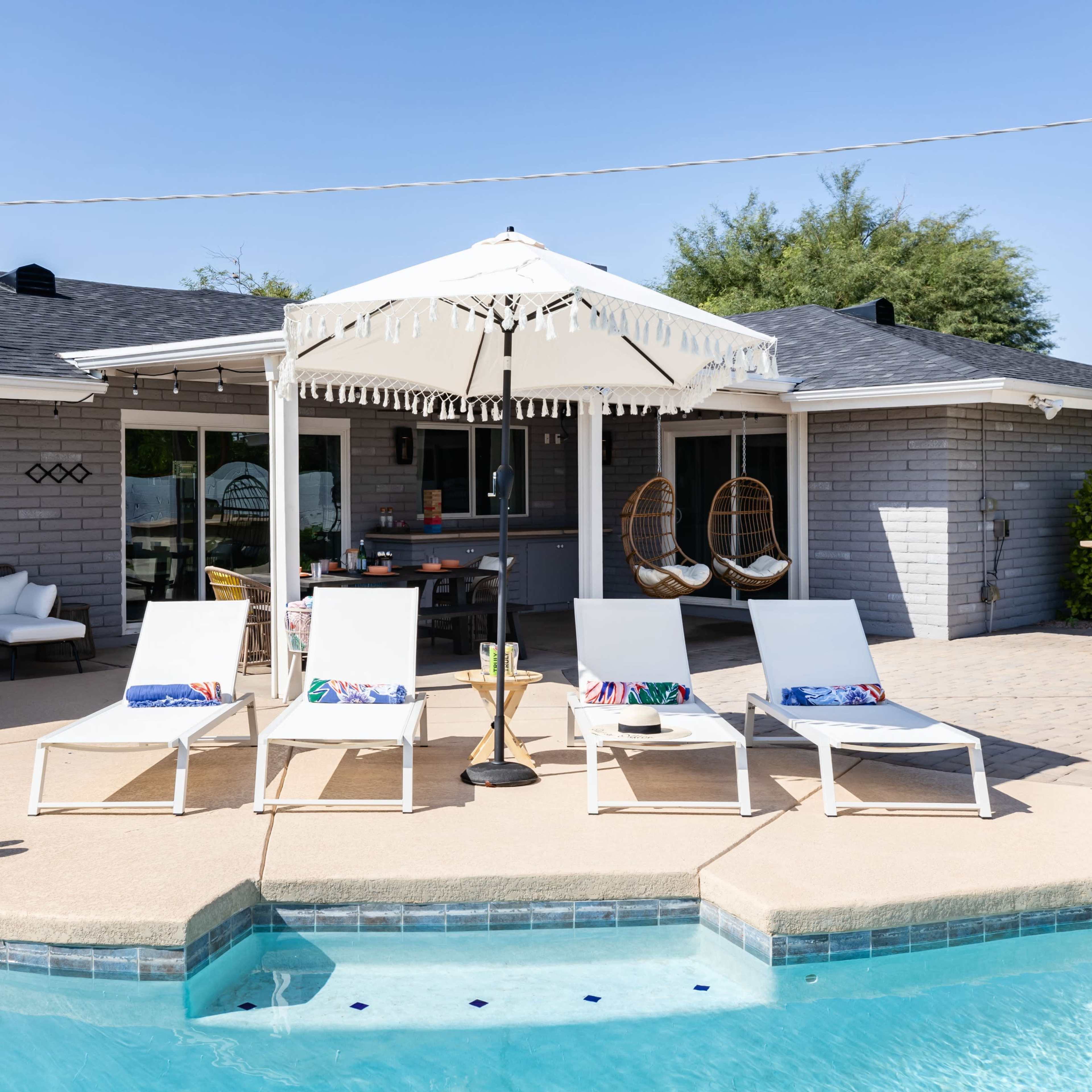 A swimming pool area features white lounge chairs and a decorative umbrella beside a modern house with a patio and palm trees in the background.