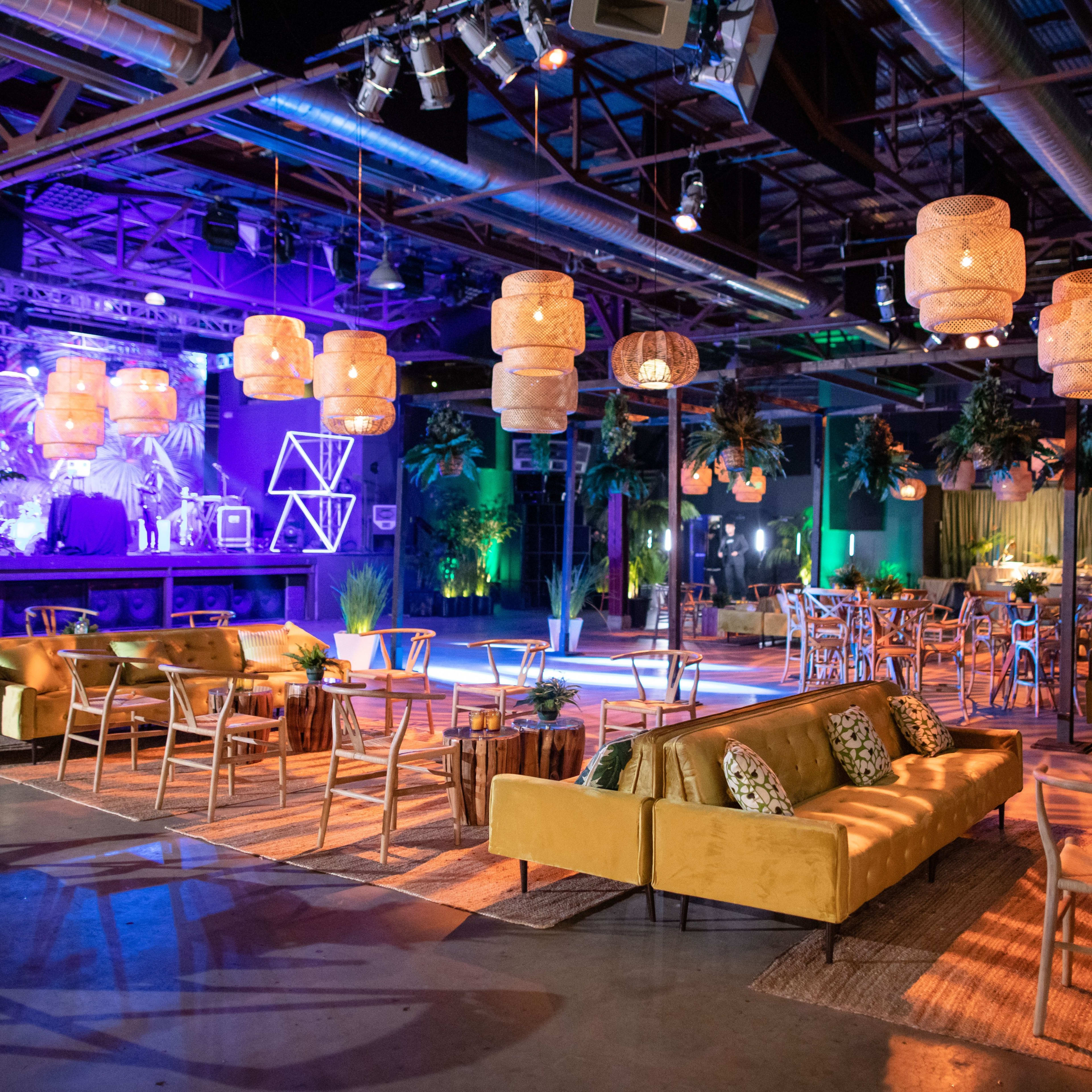 A spacious event venue features stylish seating, decorative lighting, and greenery, creating a vibrant atmosphere for gatherings.