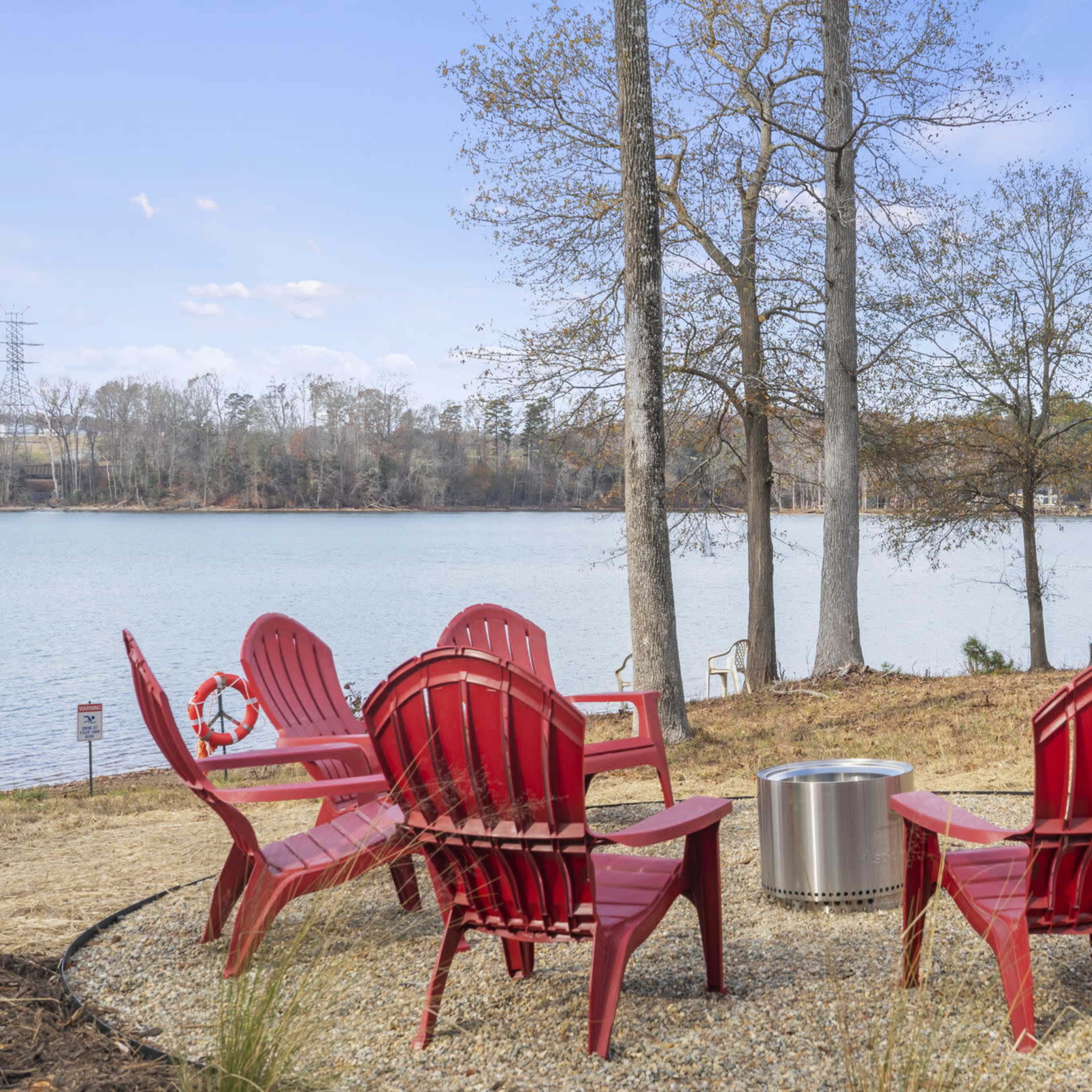 A circle of six red Adirondack chairs surrounds a metal fire pit near a calm lake, with trees in the background.