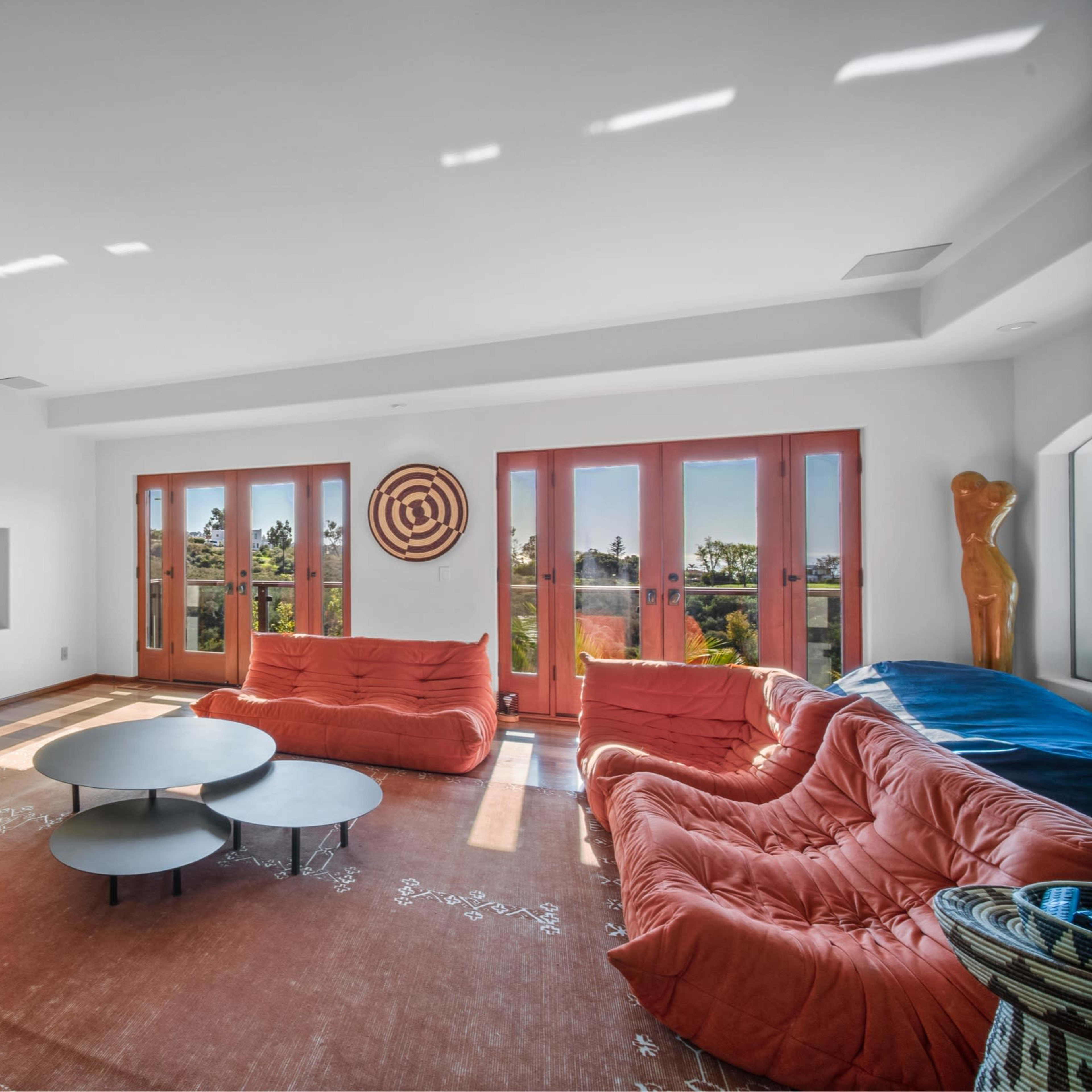 A modern living room features two orange sofas, a round coffee table, a fireplace, and large windows overlooking an outdoor view.