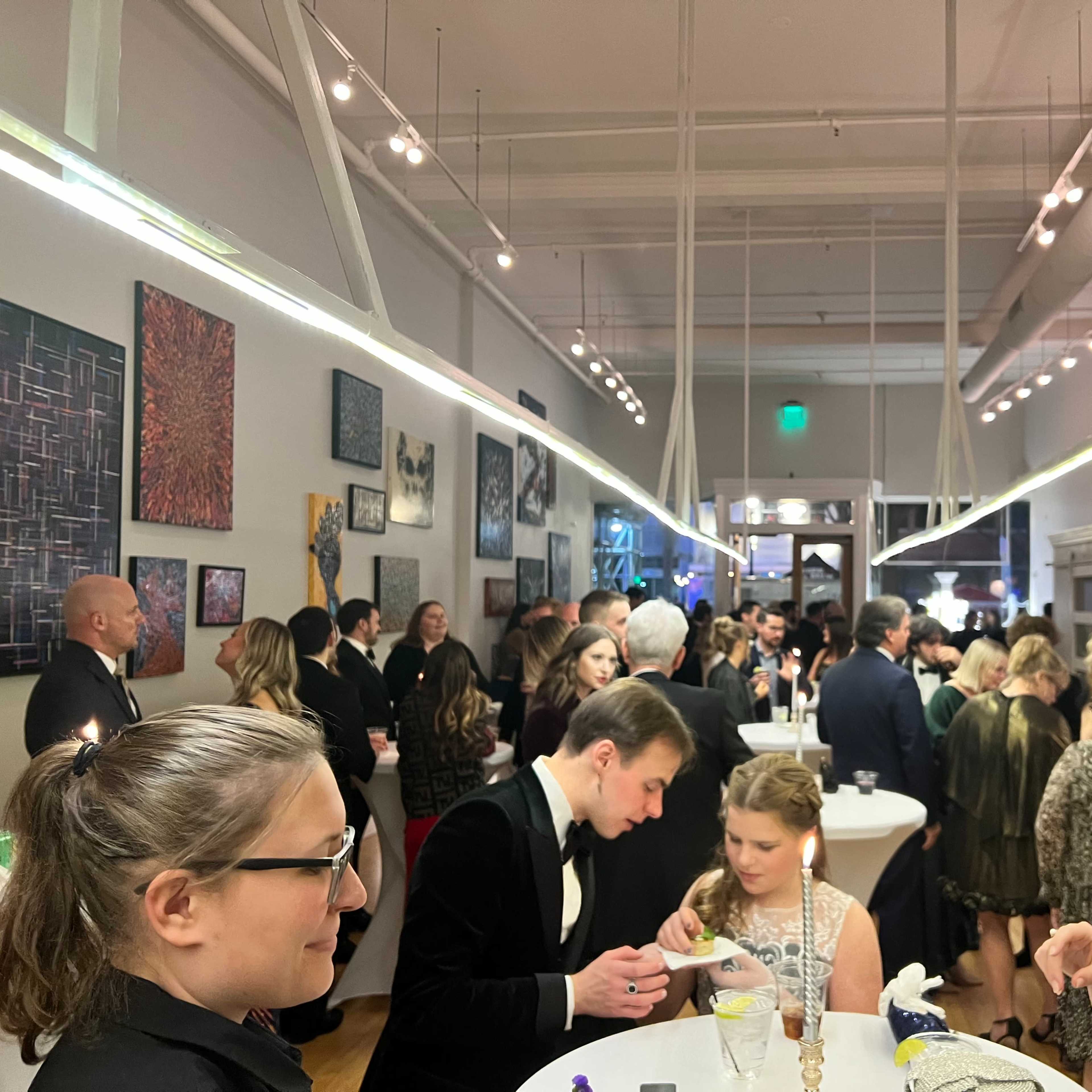 A crowd of elegantly dressed individuals mingles at a social event in an art gallery, surrounded by colorful artworks on the walls.