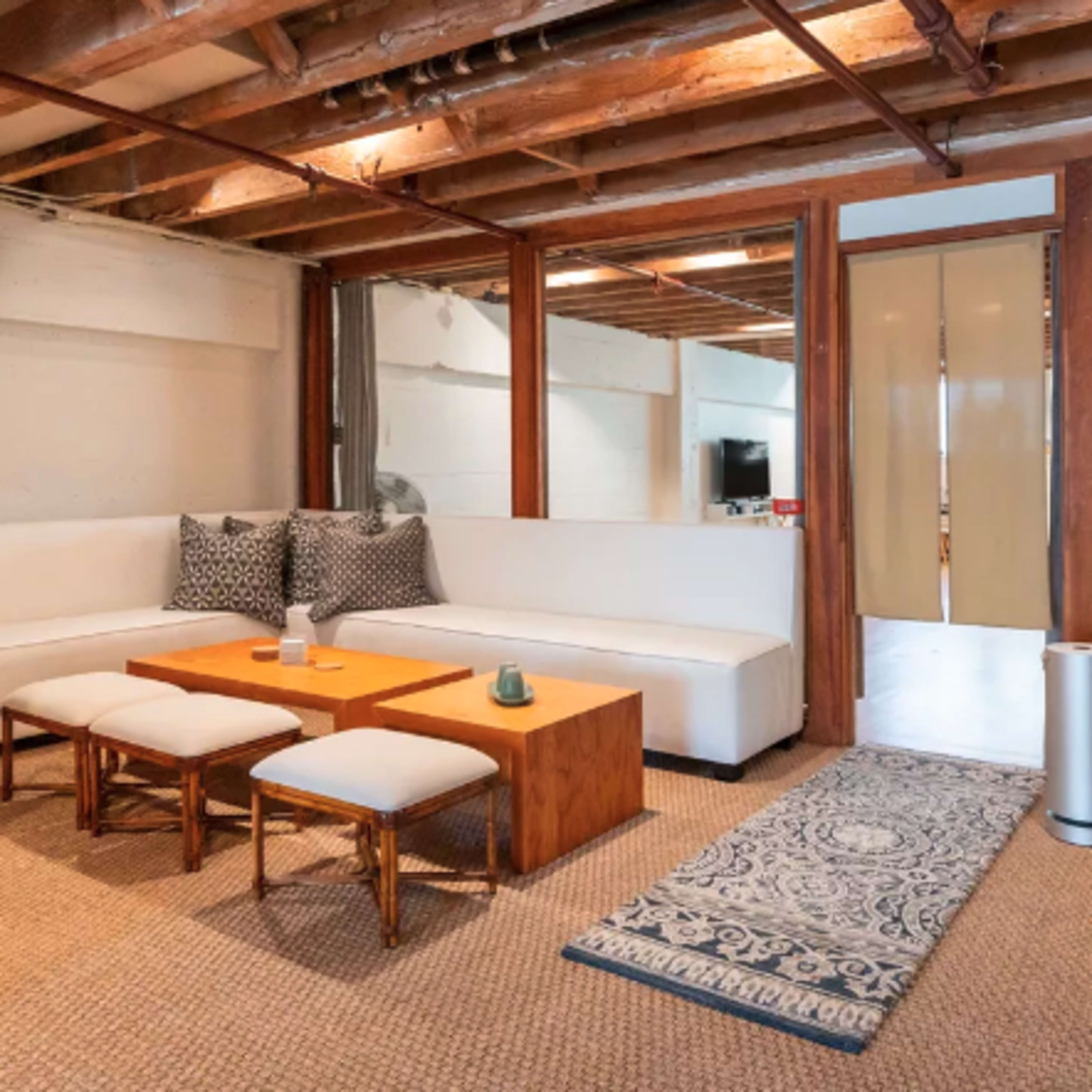 A minimalist living room with a white sectional sofa, a wooden coffee table, and patterned rugs on a textured floor.