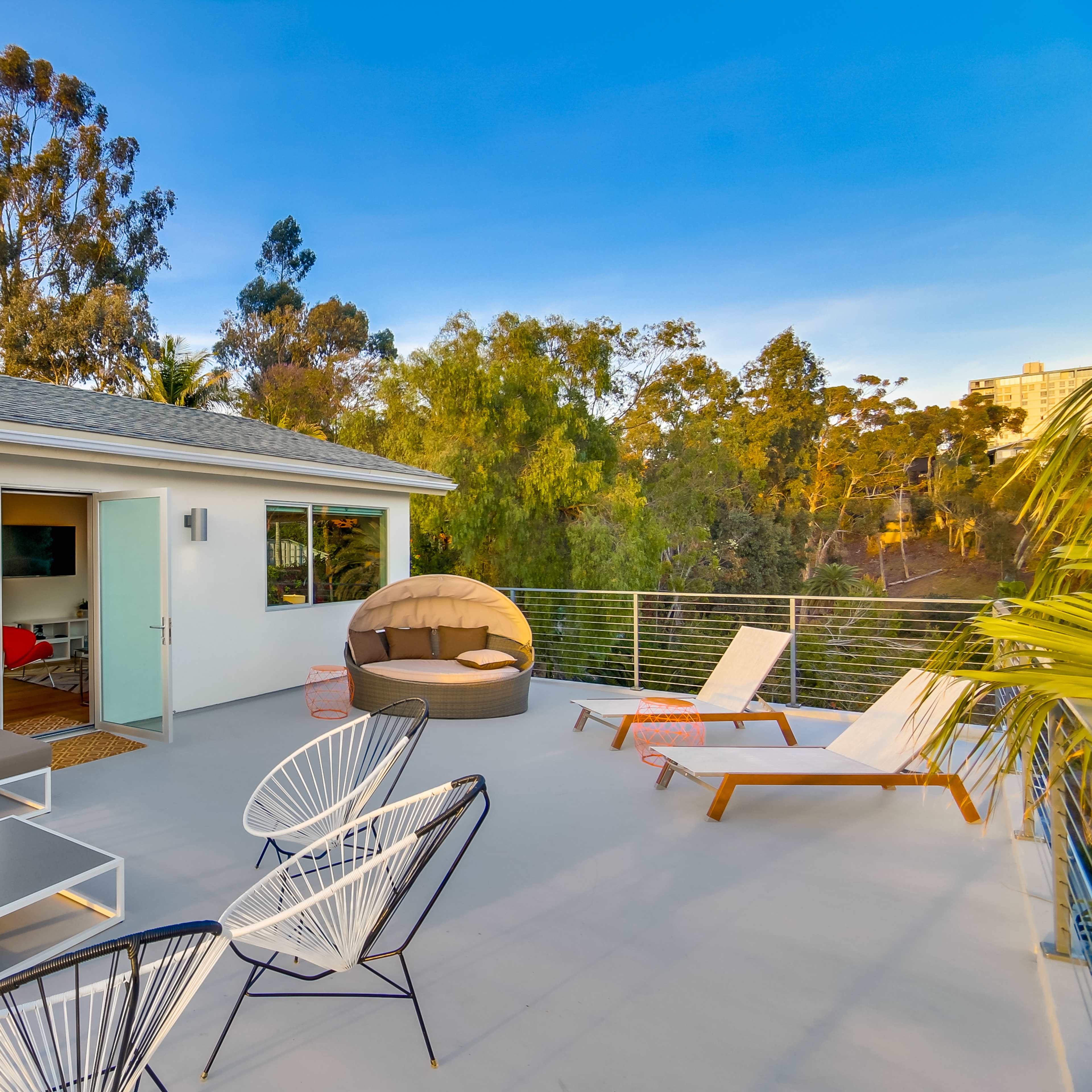 A modern rooftop terrace features a sectional sofa, lounge chairs, and a round daybed surrounded by trees and a clear sky.