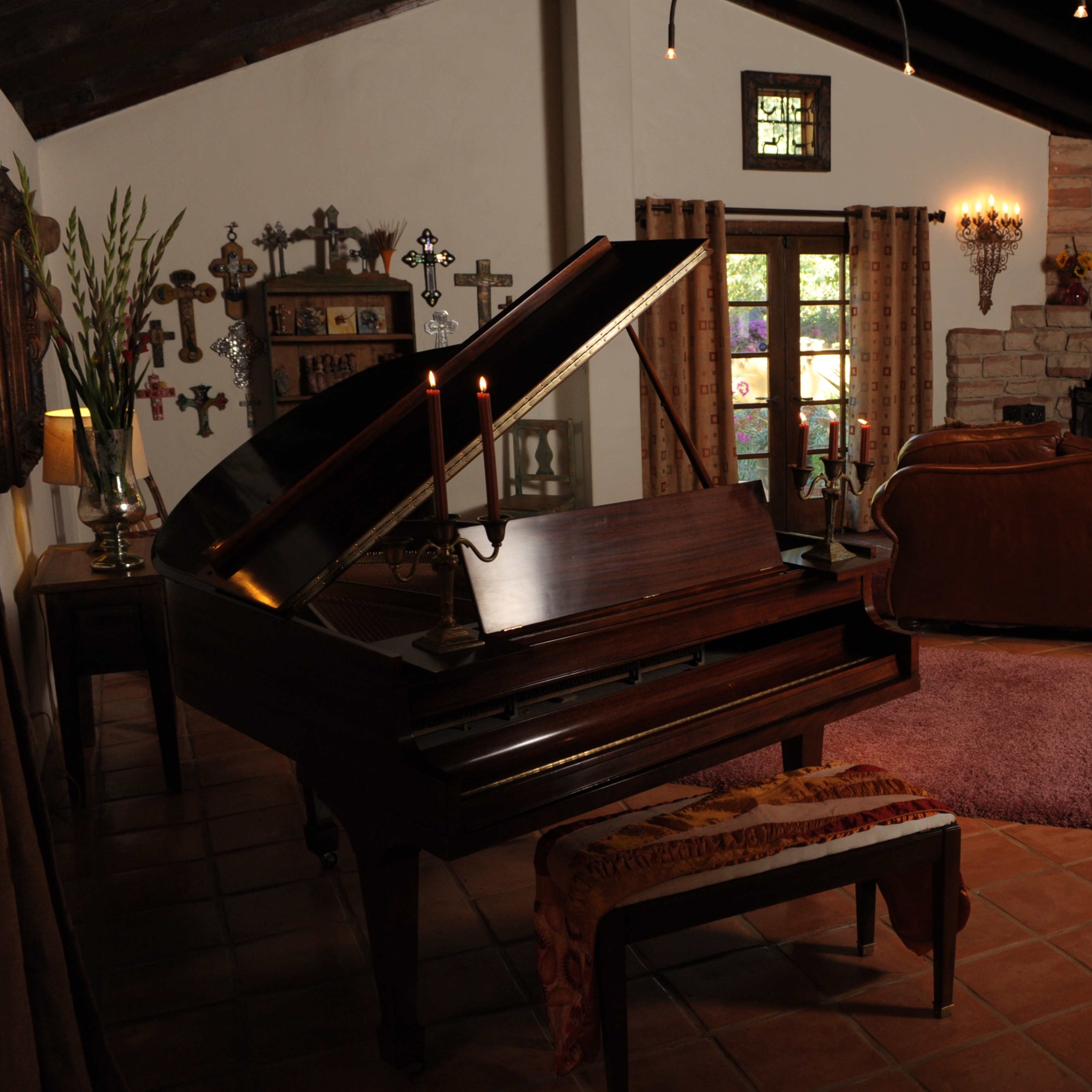 The room features a grand piano next to a brown leather sofa, with a stone fireplace and large windows letting in natural light.