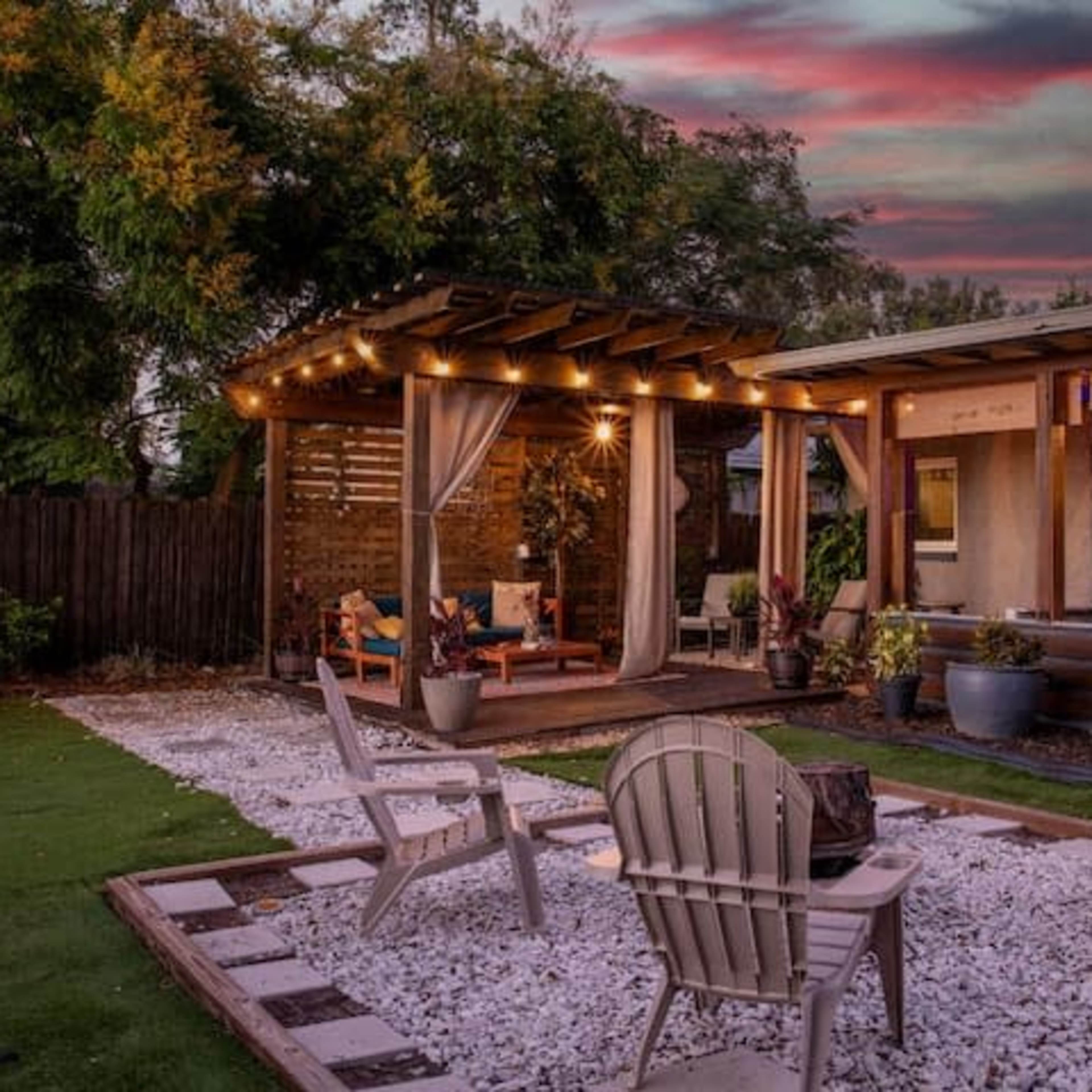 A cozy backyard features a seating area with a pergola, adorned with lights and curtains, surrounded by gravel paths and colorful plants.