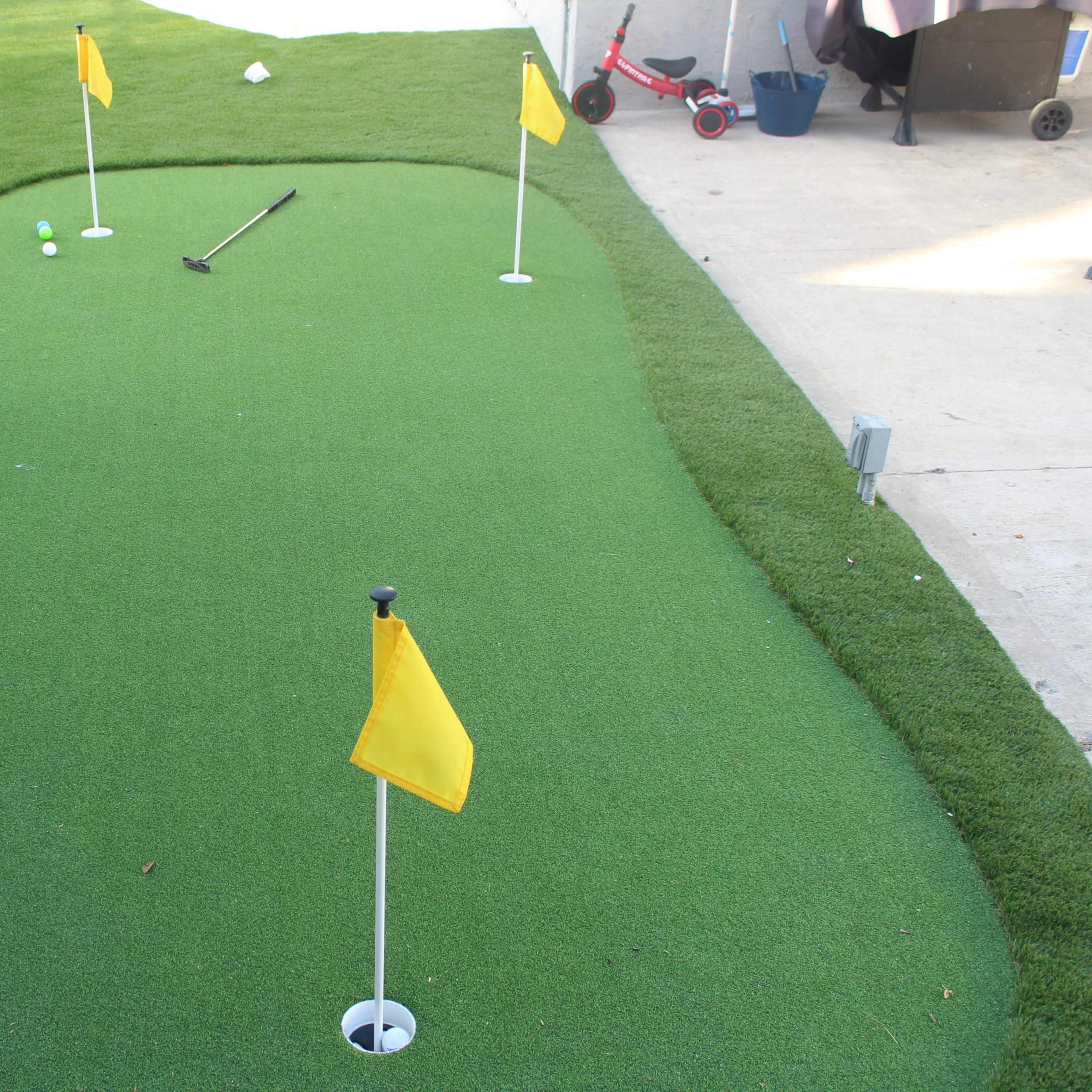 A small putting green with three yellow flags and a golf ball sits on a lawn beside a concrete patio.