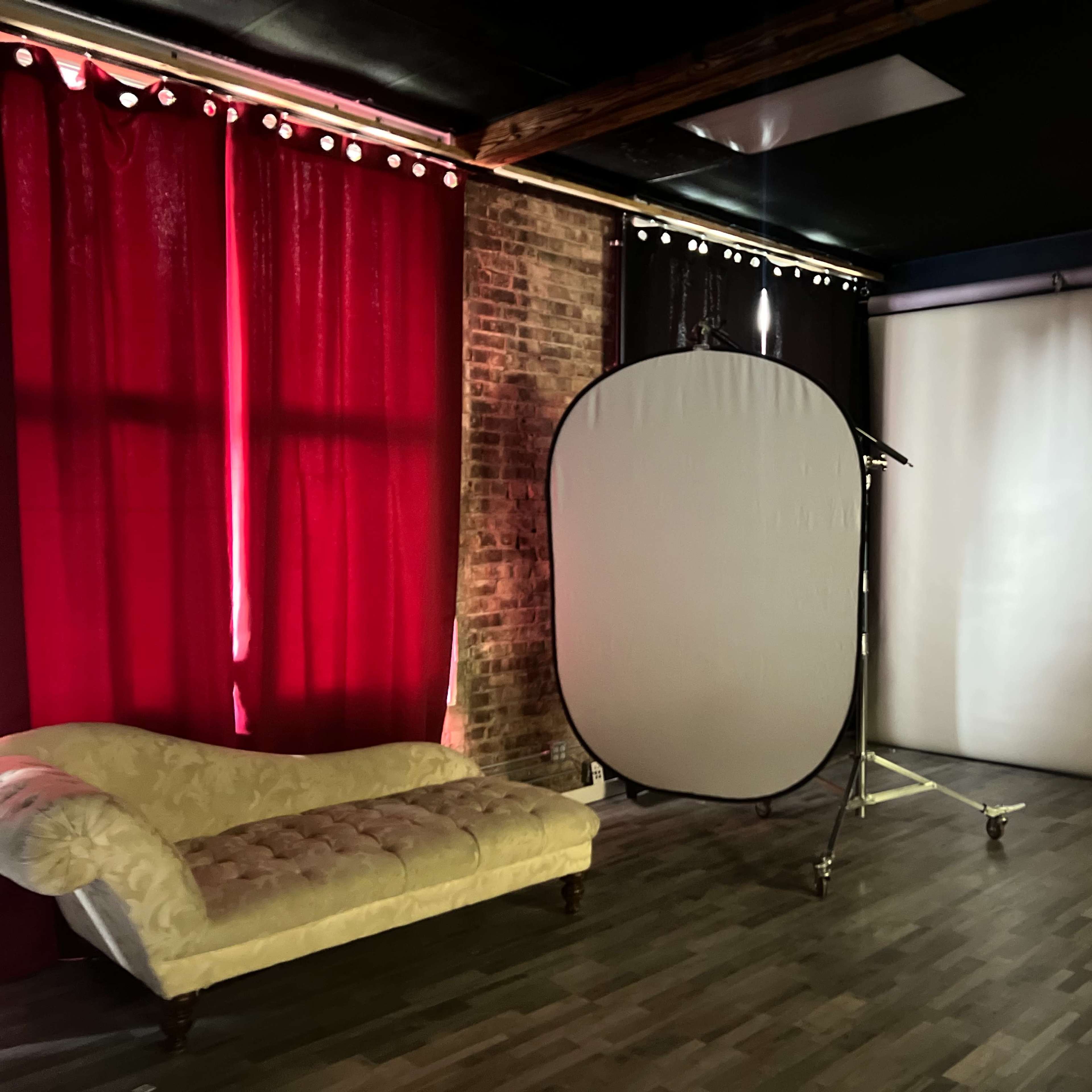 A studio space with a vintage couch, a collapsible backdrop on a stand, and two walls covered in red drapes and a plain white sheet, respectively.