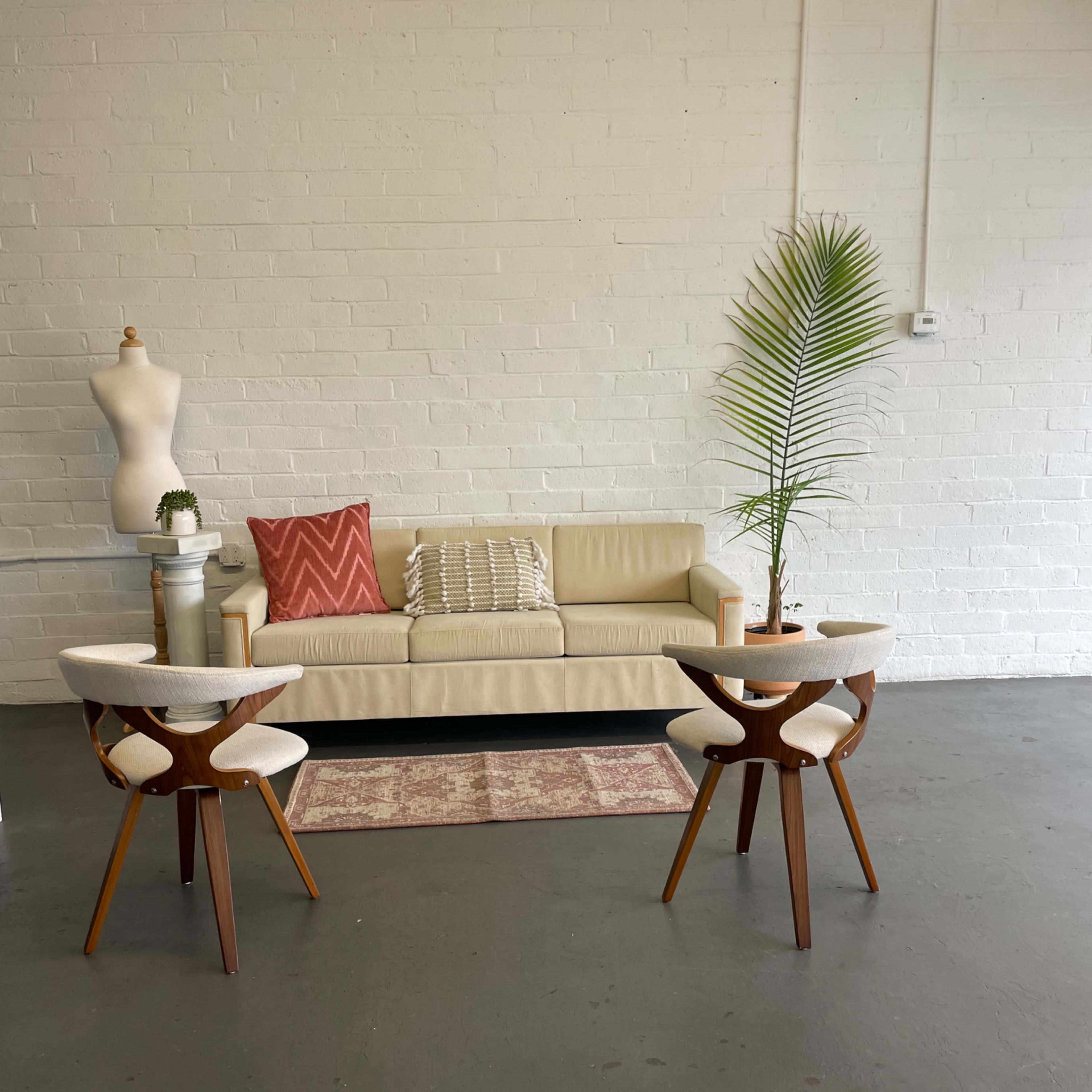 A minimalist room with a beige couch, two wooden chairs with white cushions, a small round table between them, and a decorative plant.