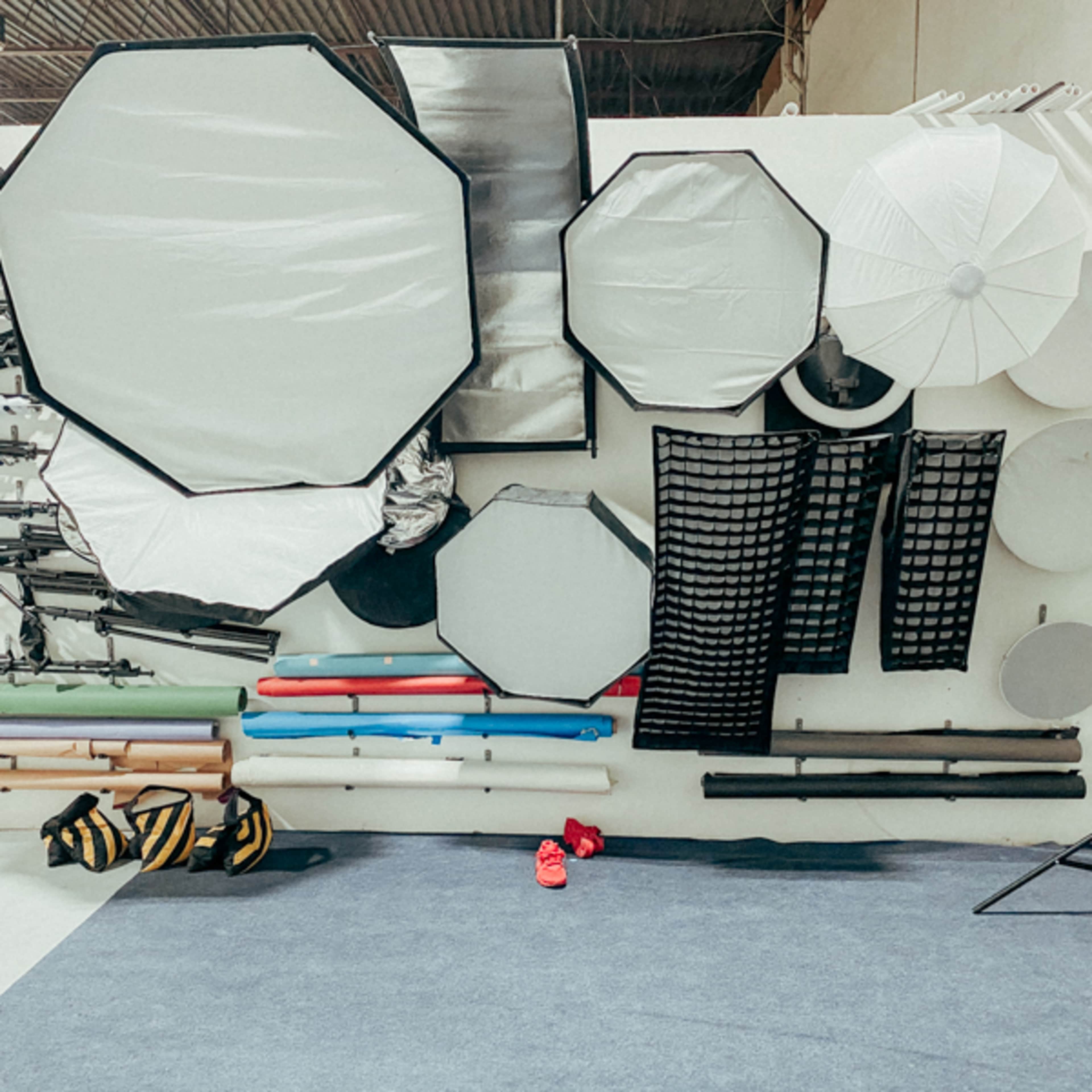 A wall in a photography studio with various lighting equipment, including umbrellas, softboxes, and reflectors, arranged alongside colorful backdrops and support stands.