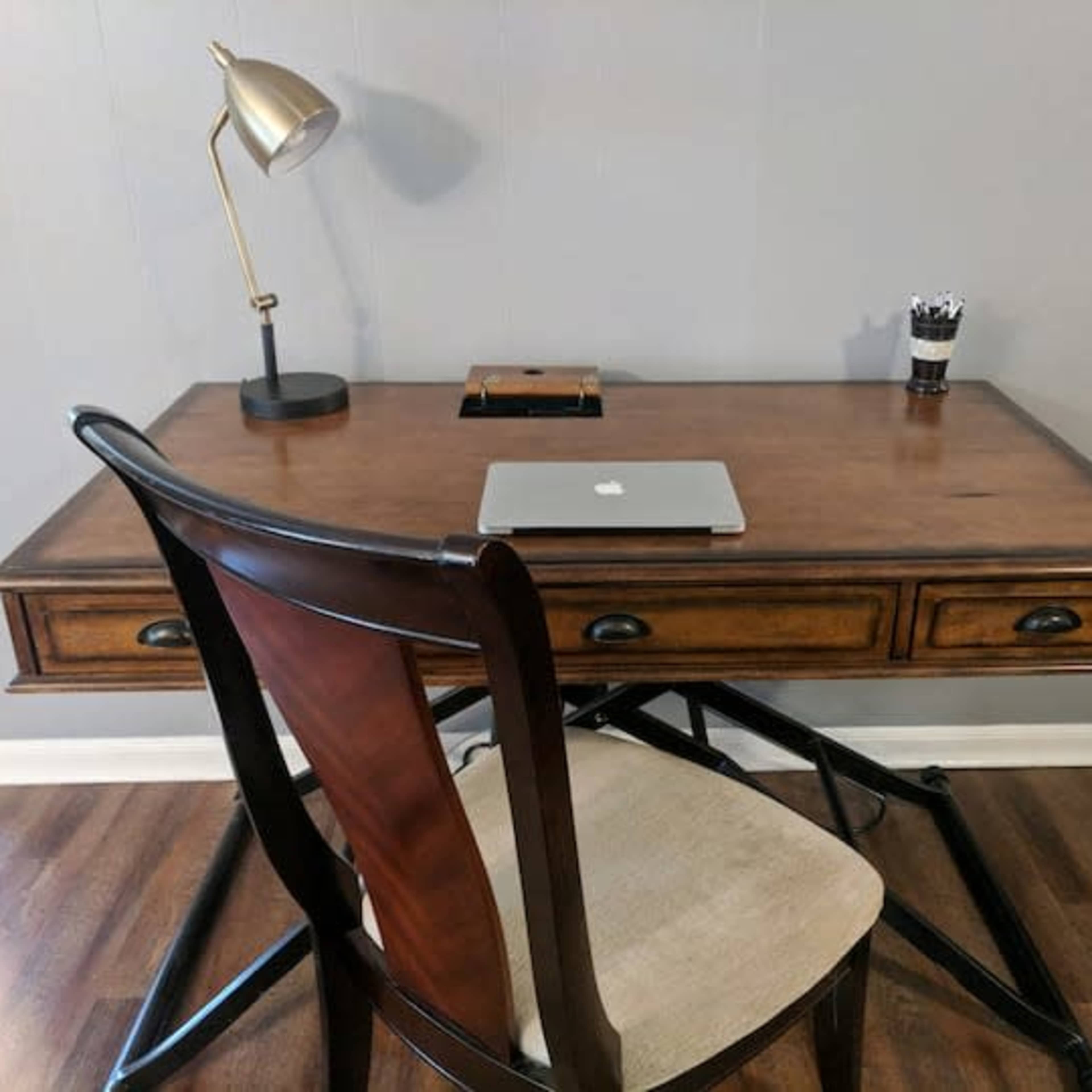 A wooden desk with a laptop, a pen holder, and a desk lamp is positioned against a gray wall.