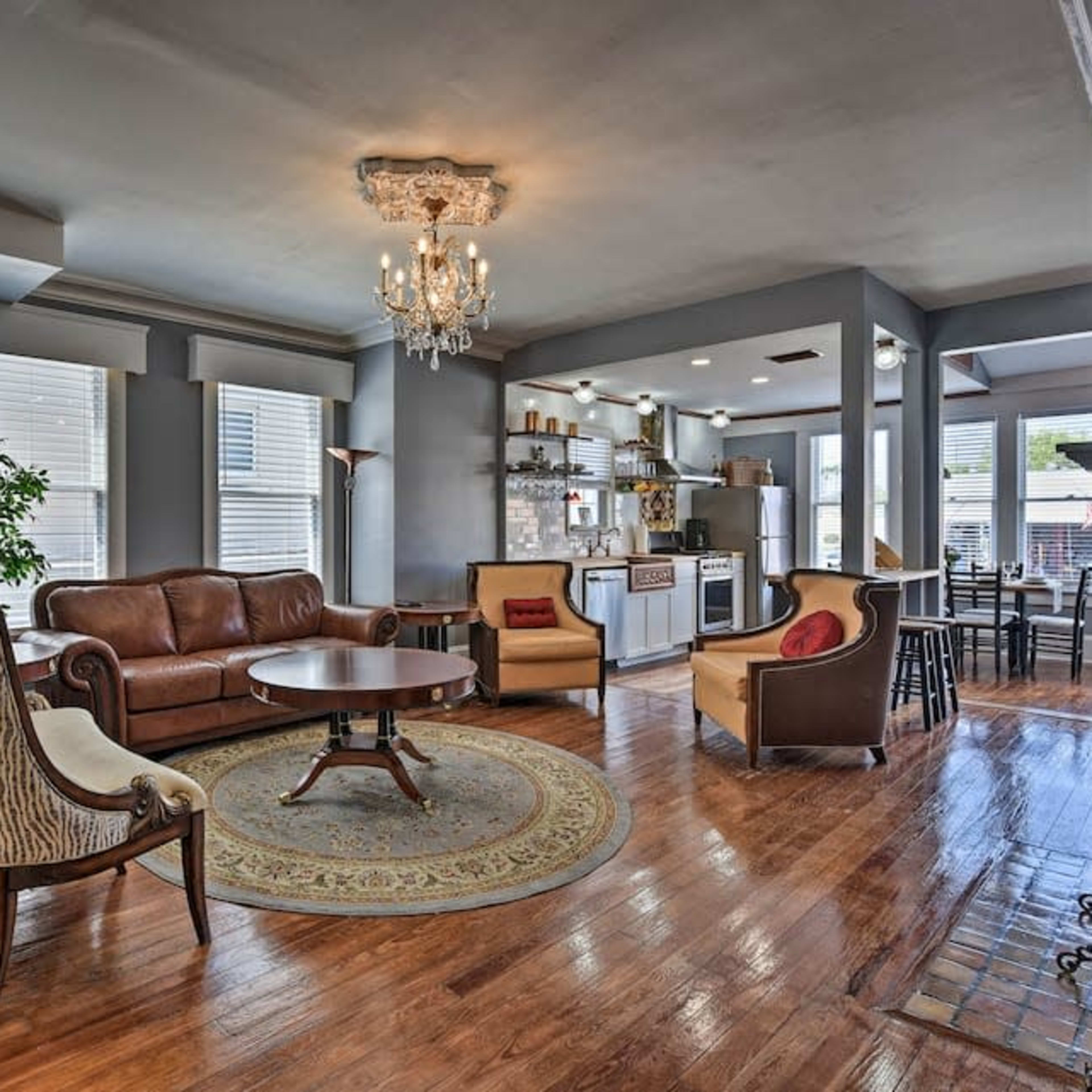 A spacious living room with hardwood floors, a brown leather sofa, and a round wooden coffee table, surrounded by windows and a kitchen area in the background.