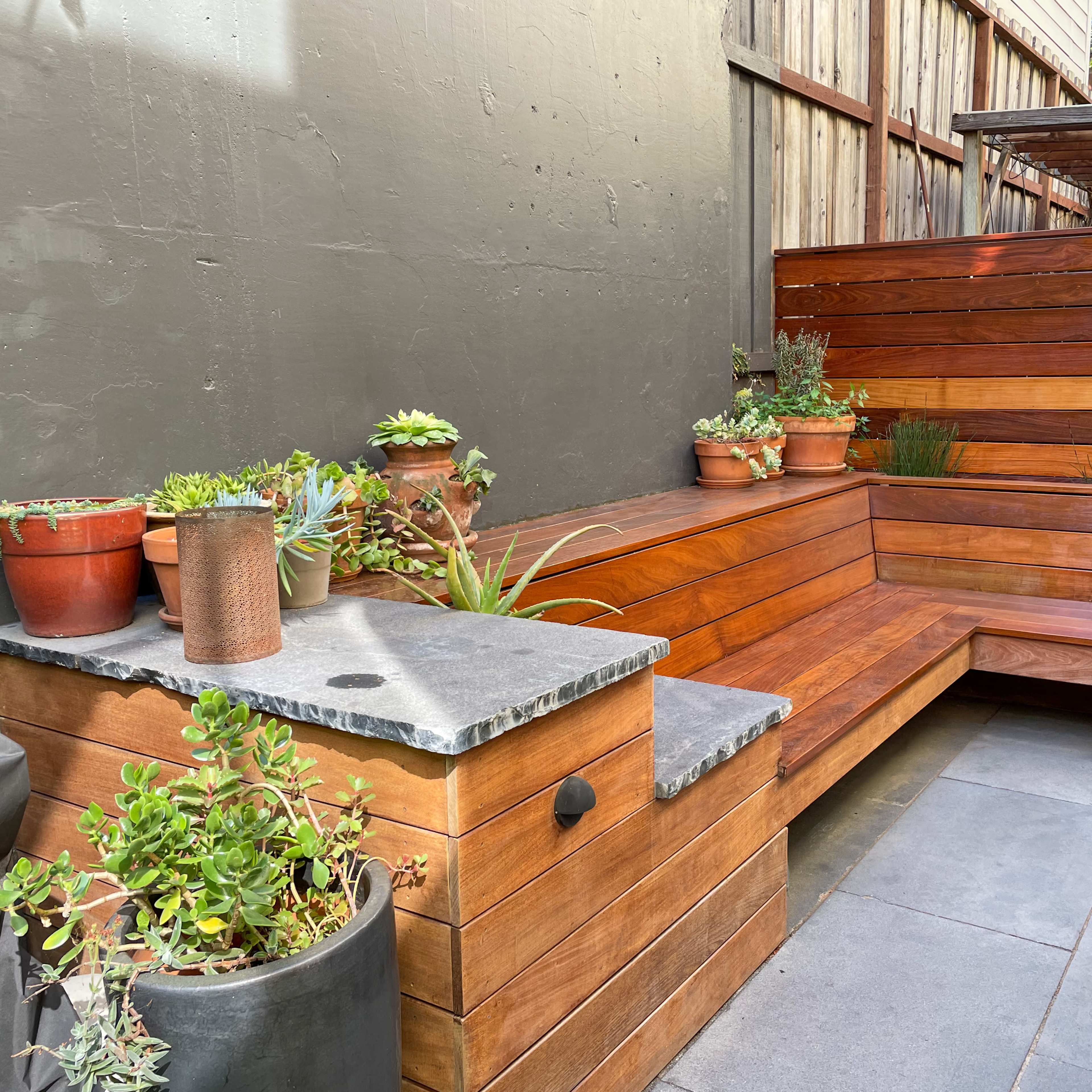 A wooden seating area bordered by potted succulents and a stone tabletop against a gray wall.