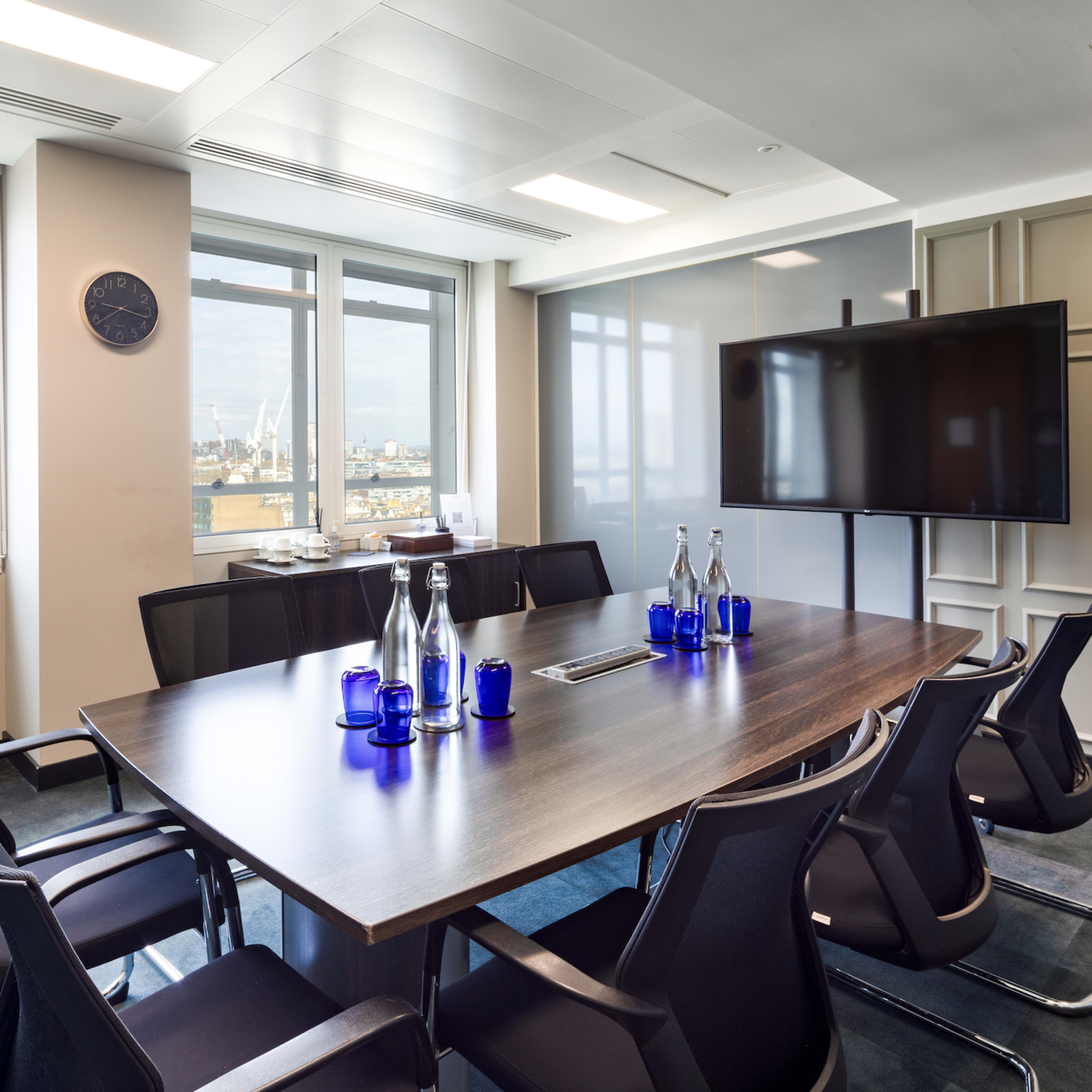 A modern conference room features a large wooden table surrounded by black chairs, with floor-to-ceiling windows offering a view of the city and a television screen mounted on the wall.