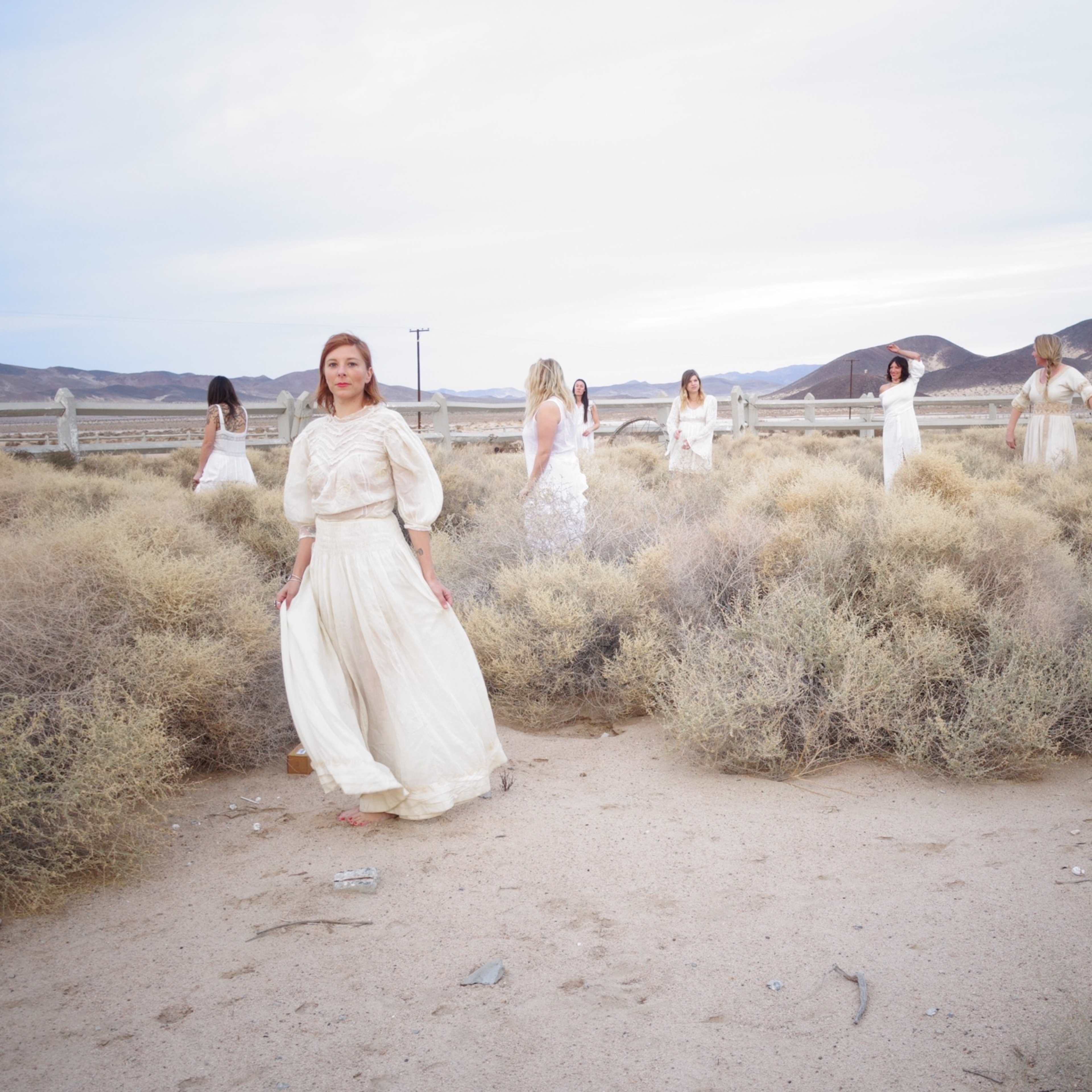 A group of women in white dresses stands scattered among dry shrubs in a desert landscape with distant mountains.
