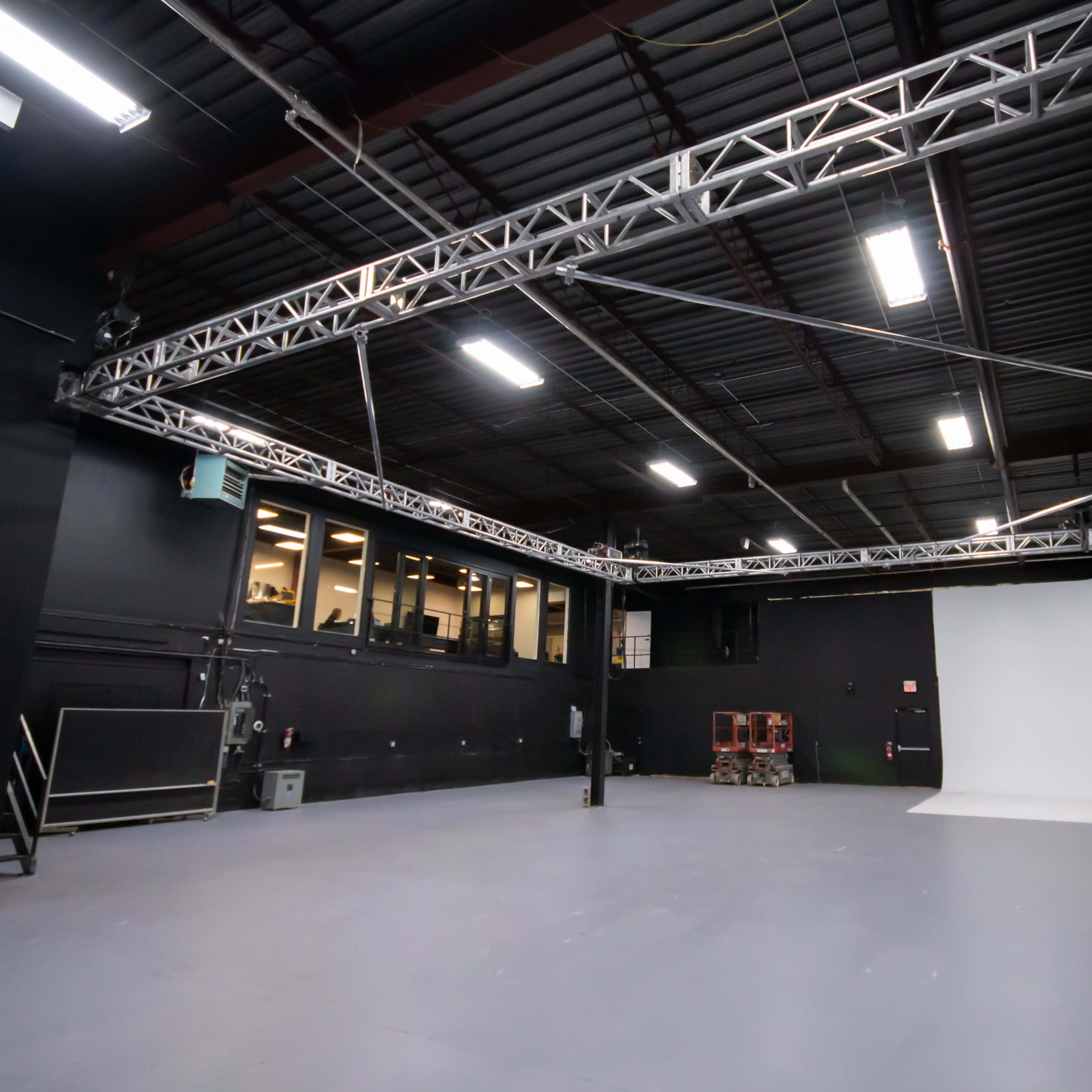 An empty photography studio with black walls, a gray floor, and a white backdrop at one end.
