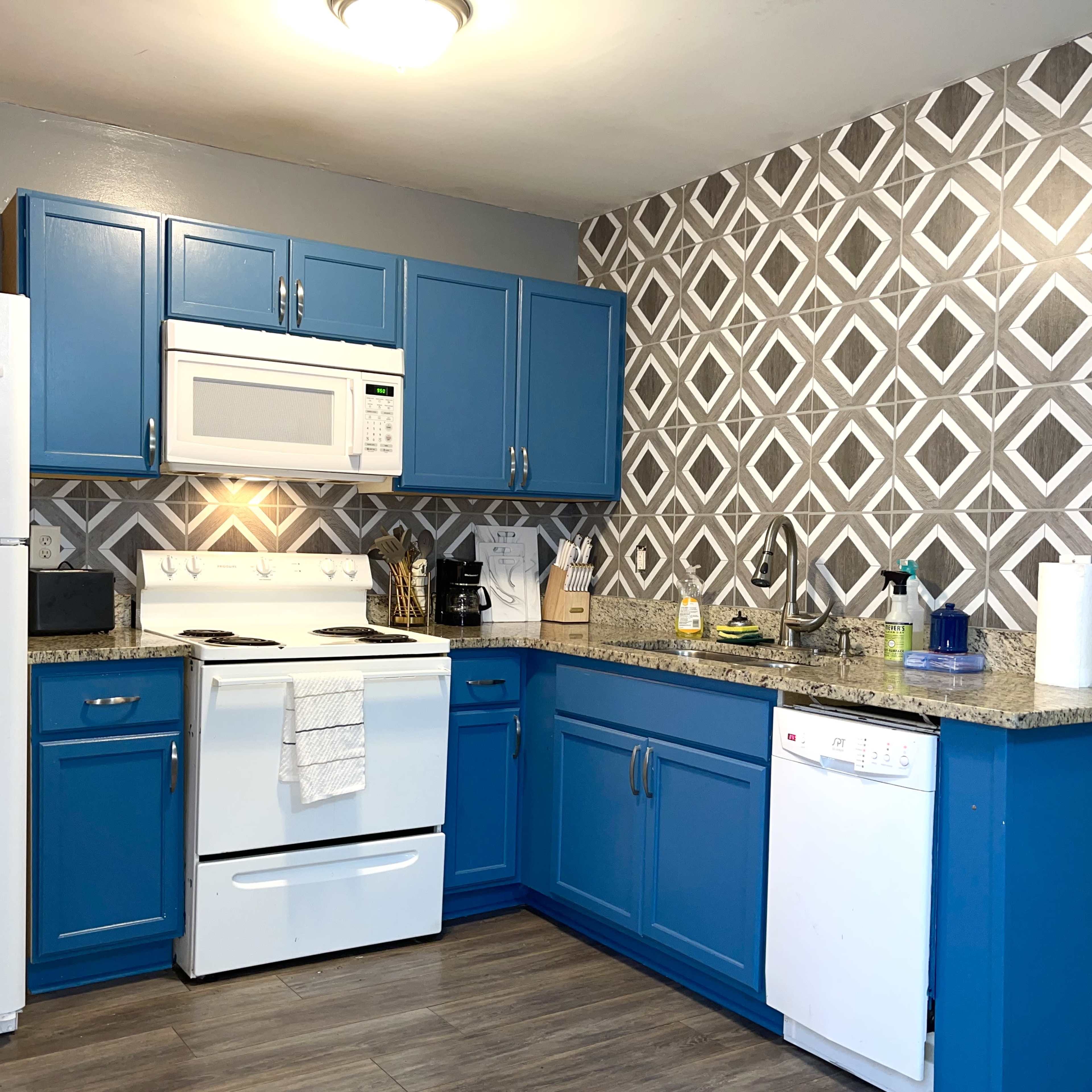 A kitchen with blue cabinets, a white stove and microwave, a refrigerator, and a patterned gray and white backsplash.