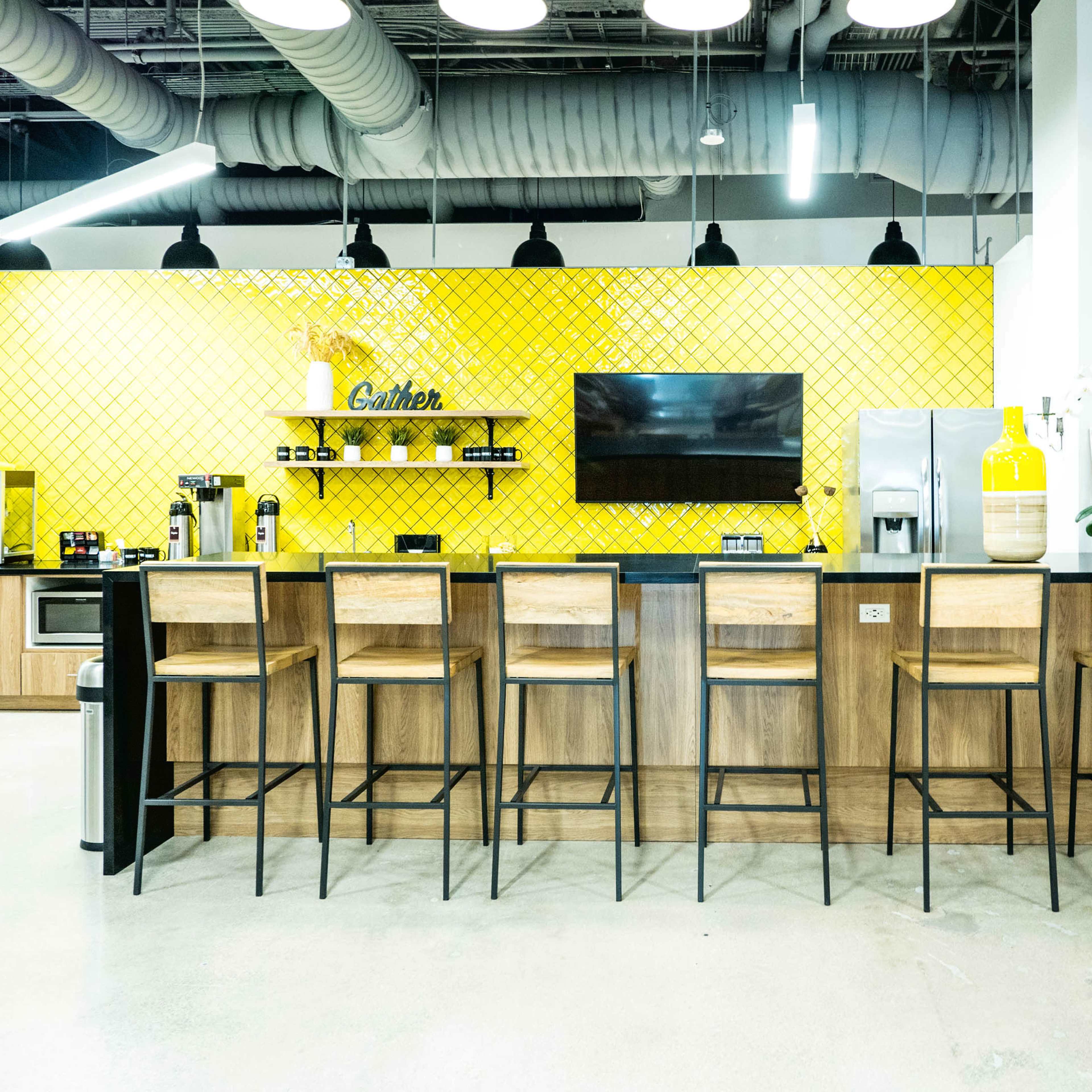 A modern kitchen with a yellow tiled wall, a large screen, and an island bar with high wooden stools arranged in front.