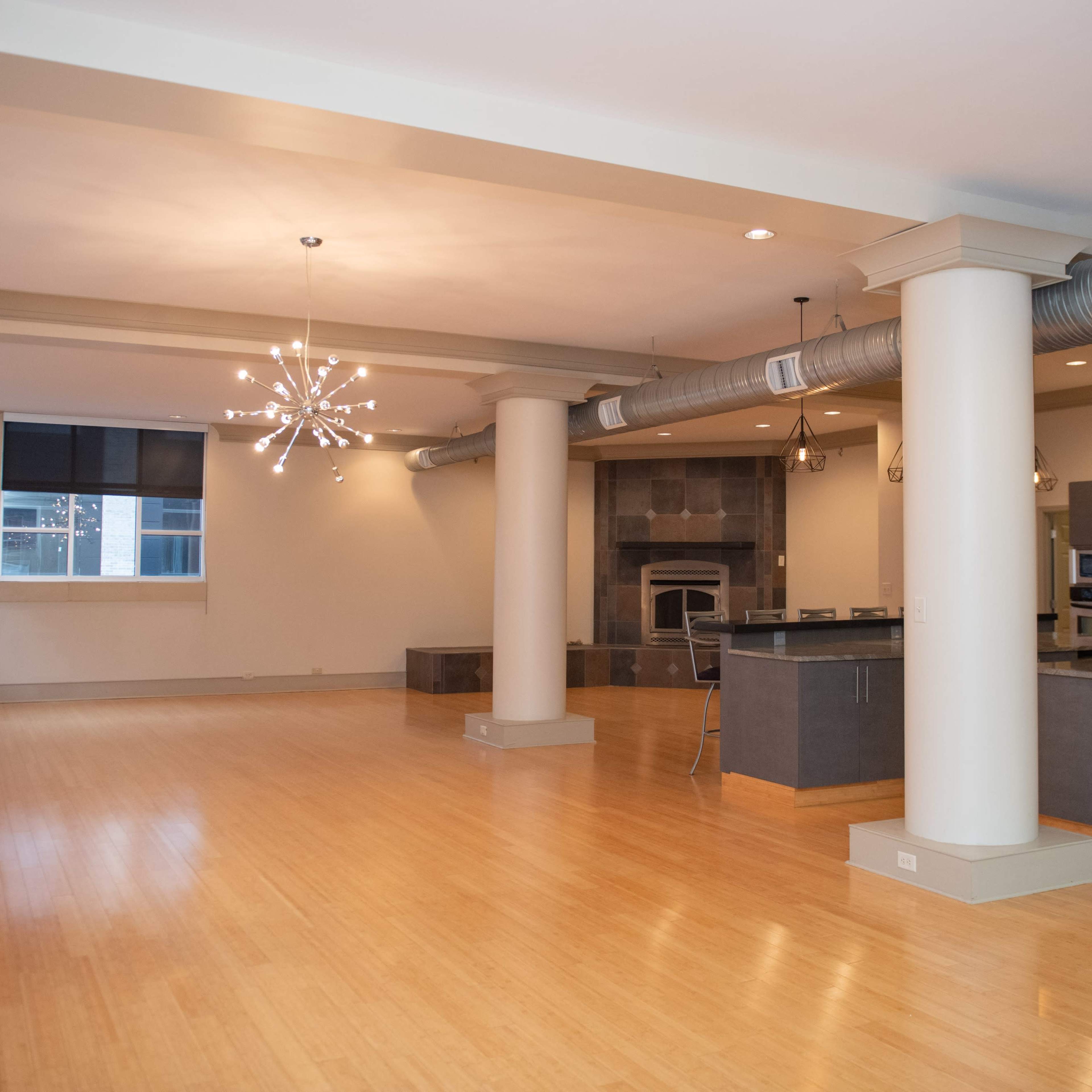 A spacious, modern open-concept living area with wooden flooring, large windows, and a kitchen featuring dark cabinetry and stainless steel appliances.