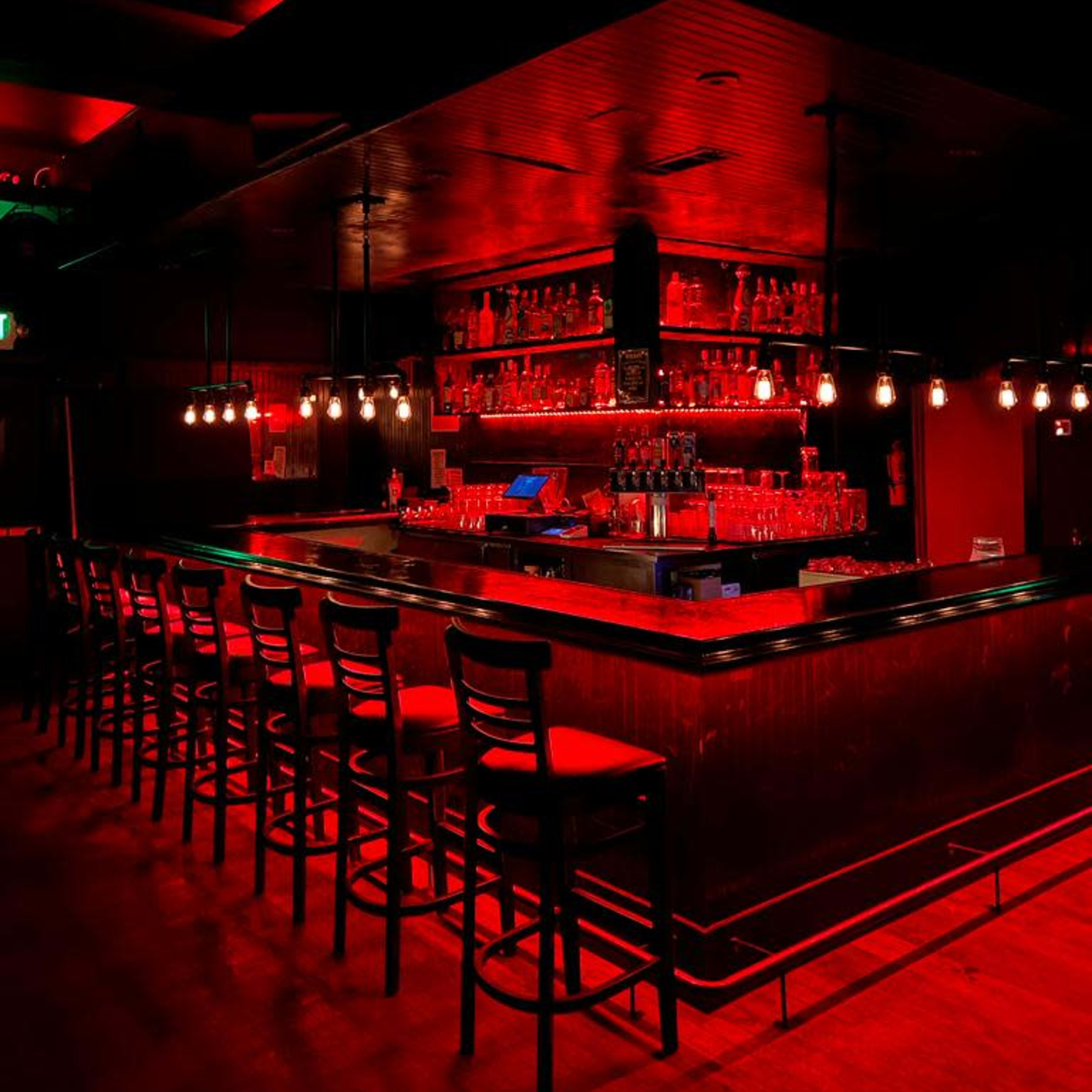 A dimly lit bar with red lighting and a long counter lined with empty chairs.