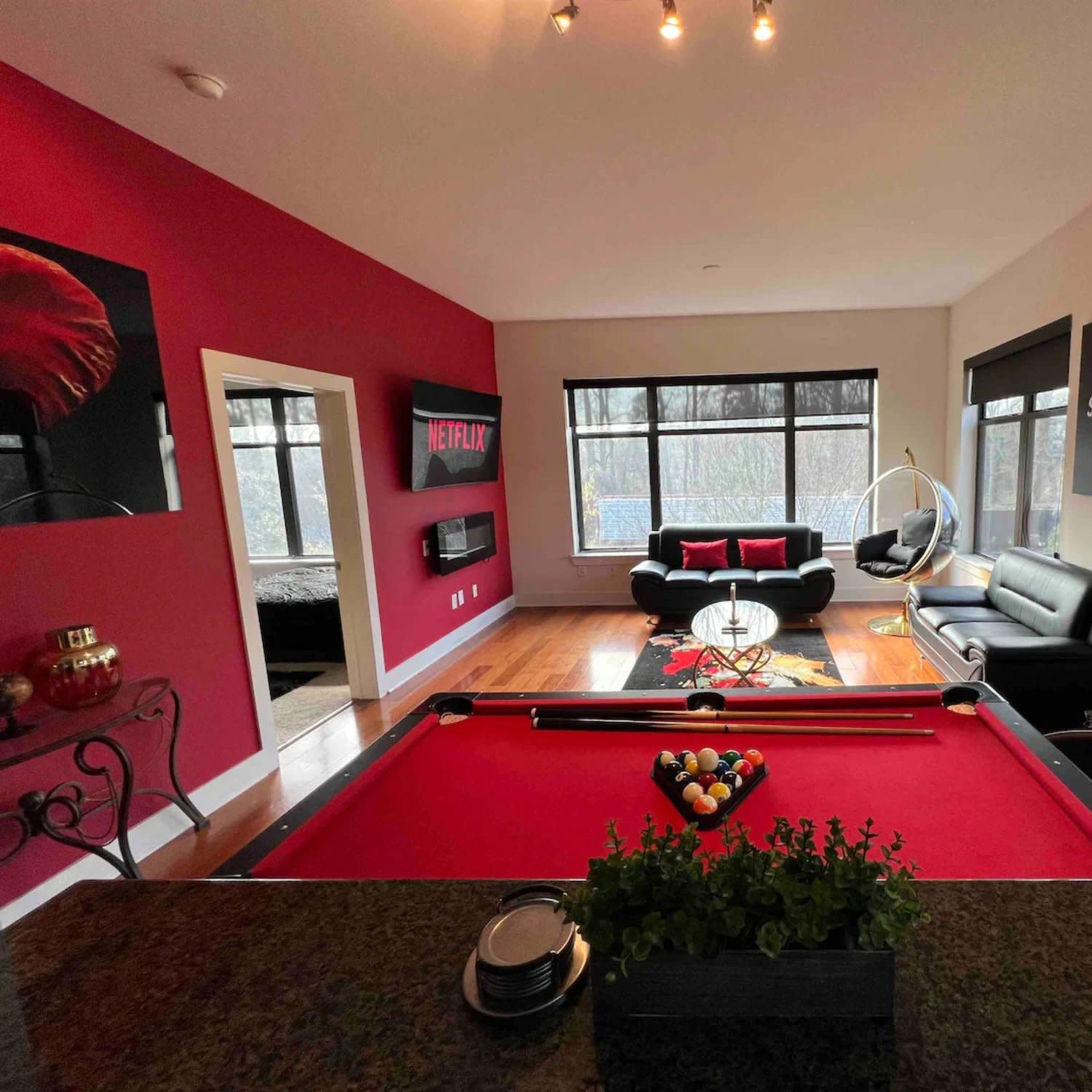 A modern living space featuring a pool table at the forefront, a seating area with black sofas, and large windows providing natural light.