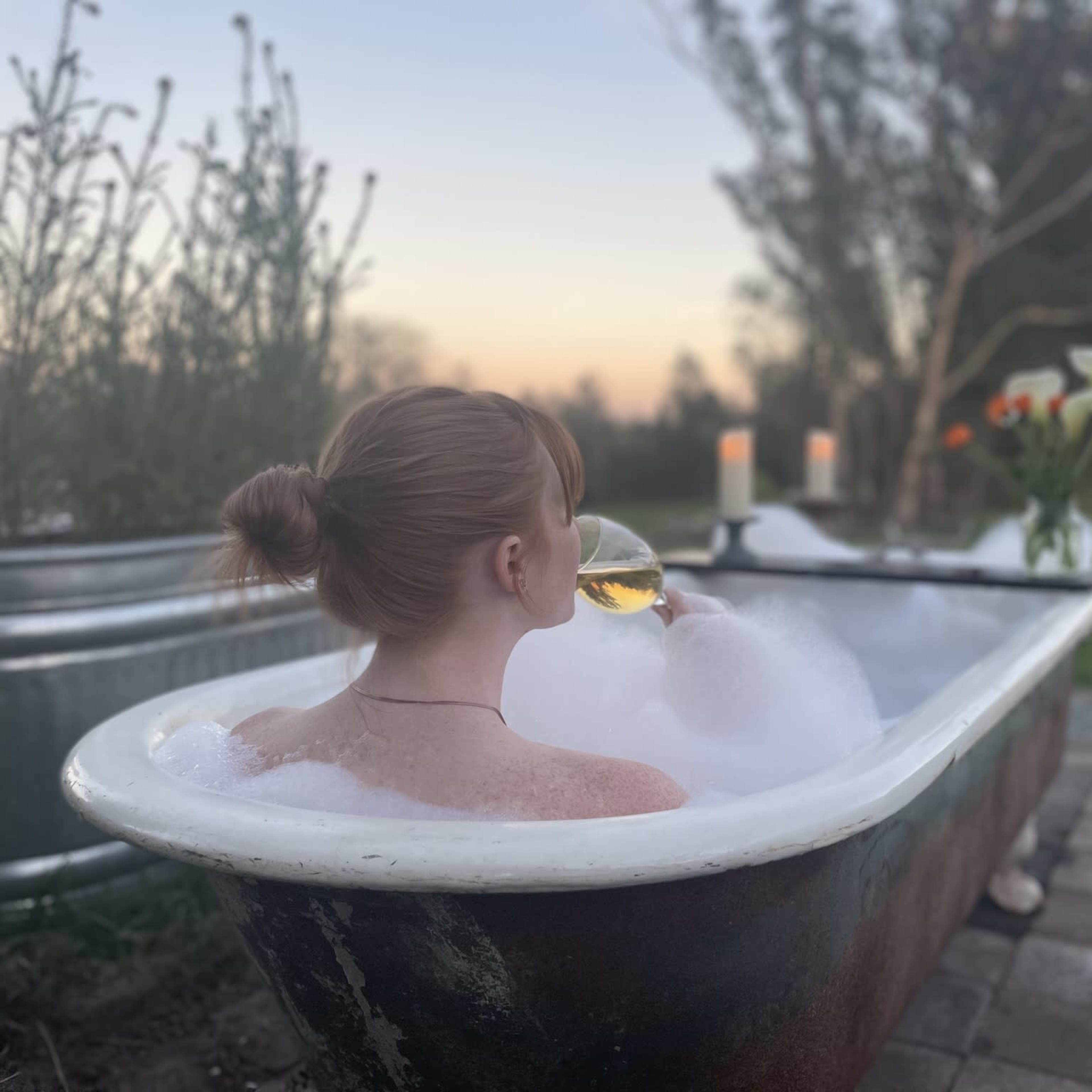 A person is relaxing in a bubble-filled vintage bathtub outdoors while holding a drink, surrounded by flowers and candles at sunset.