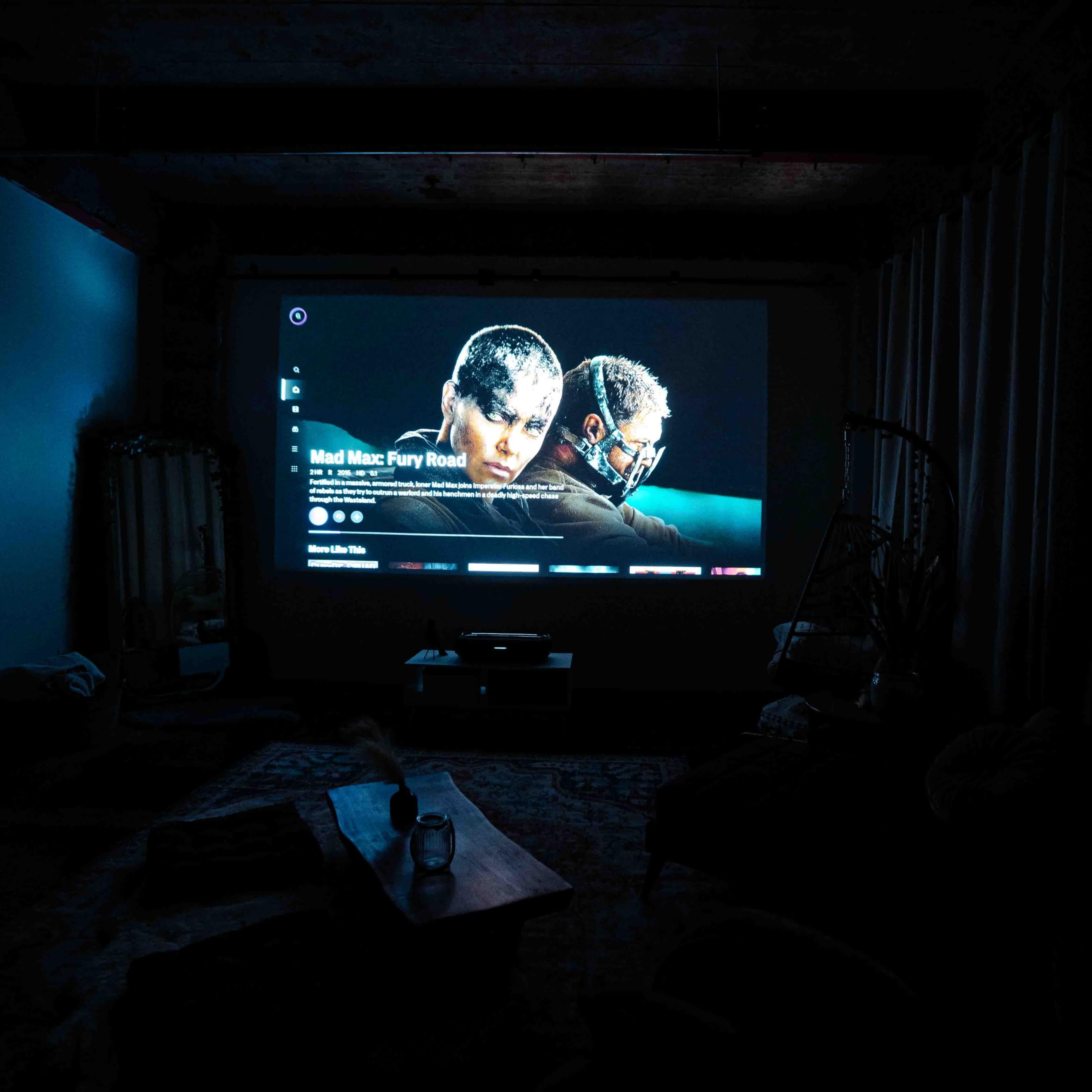 A dark room features a projector displaying the title screen for "Mad Max: Fury Road" on a wall.