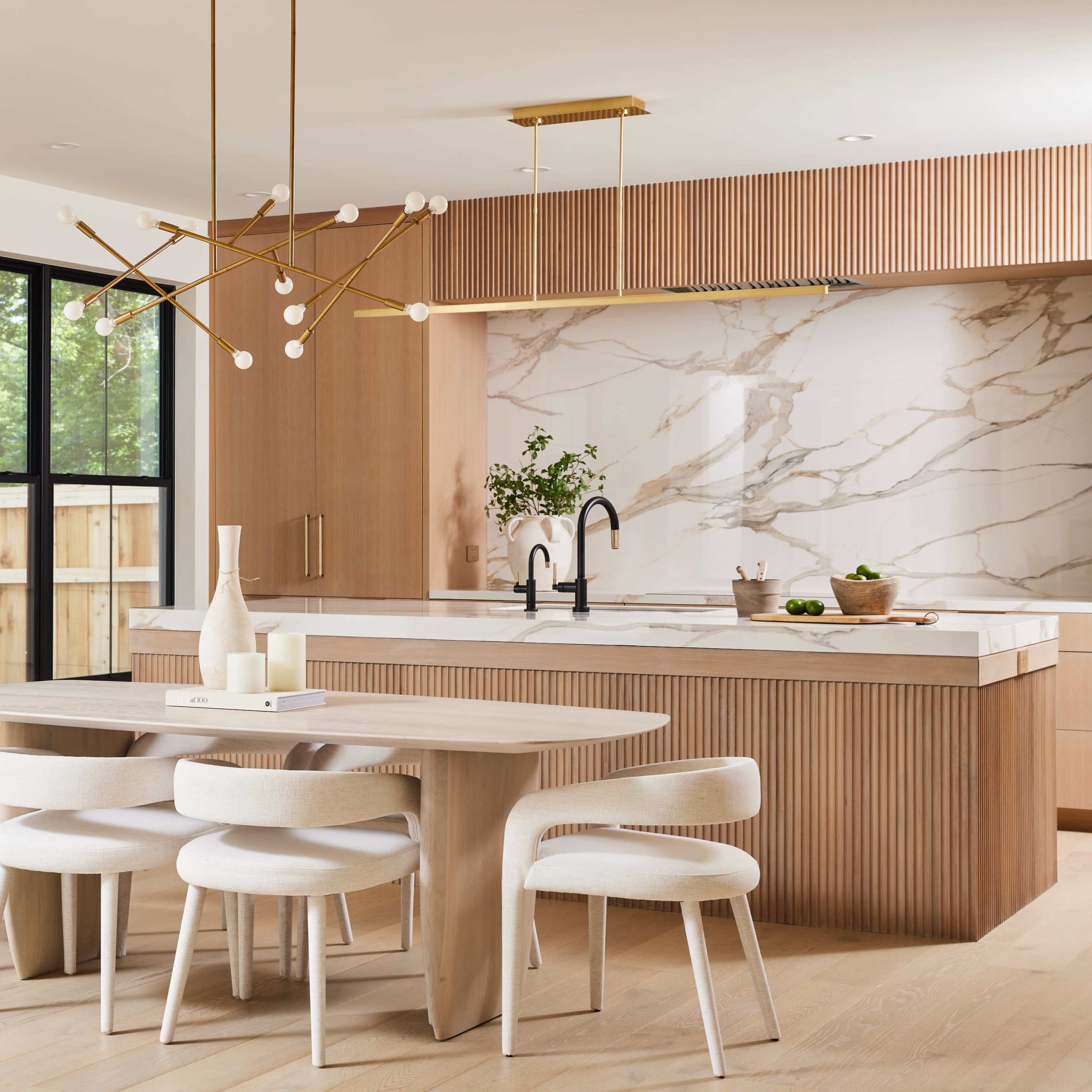 A modern kitchen features a wooden dining table surrounded by upholstered chairs, a minimalist chandelier, and a marble accent wall with a spacious island.