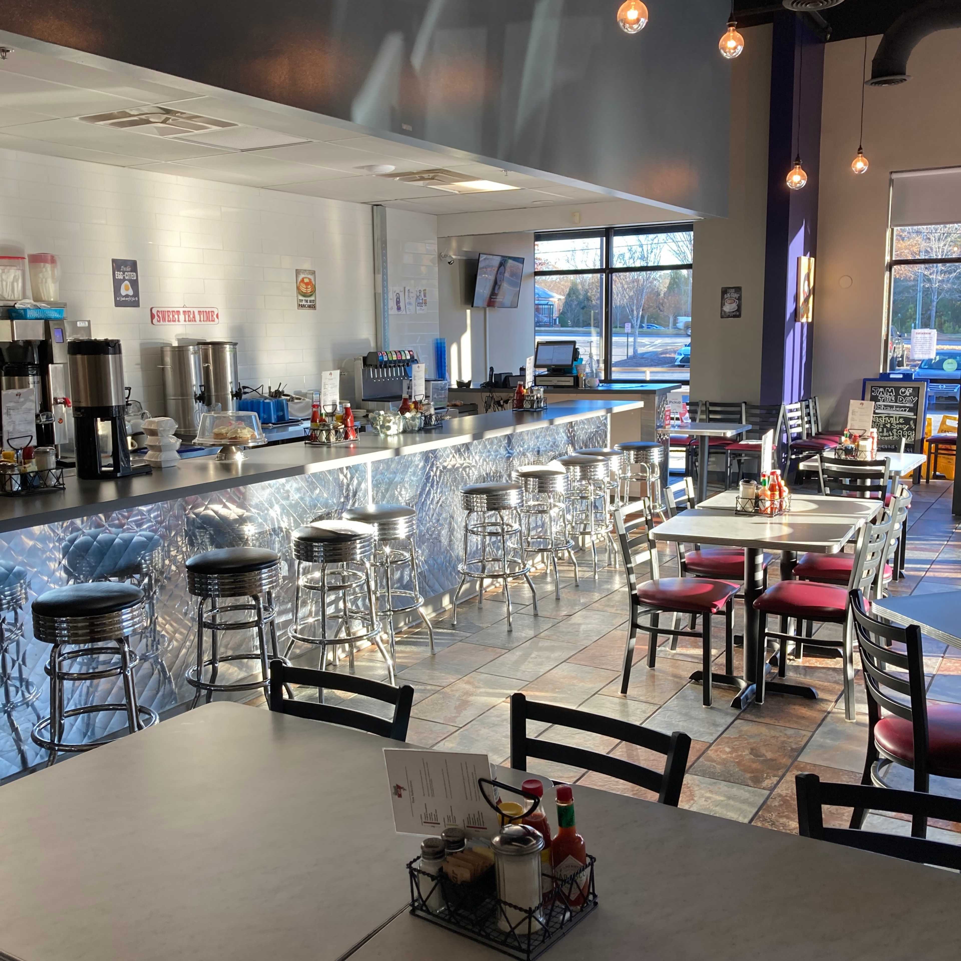 A modern diner interior with metal stools and tables, featuring a counter with drink stations and condiments visible on the tables.